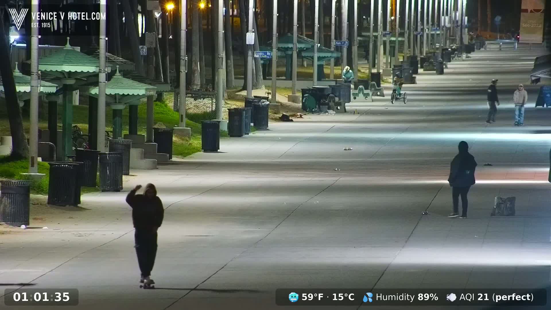 A nighttime, well-lit paved walkway, lined with trash cans and park benches under palm trees, shows several people walking and one person skateboarding, with a clear sky and high humidity.