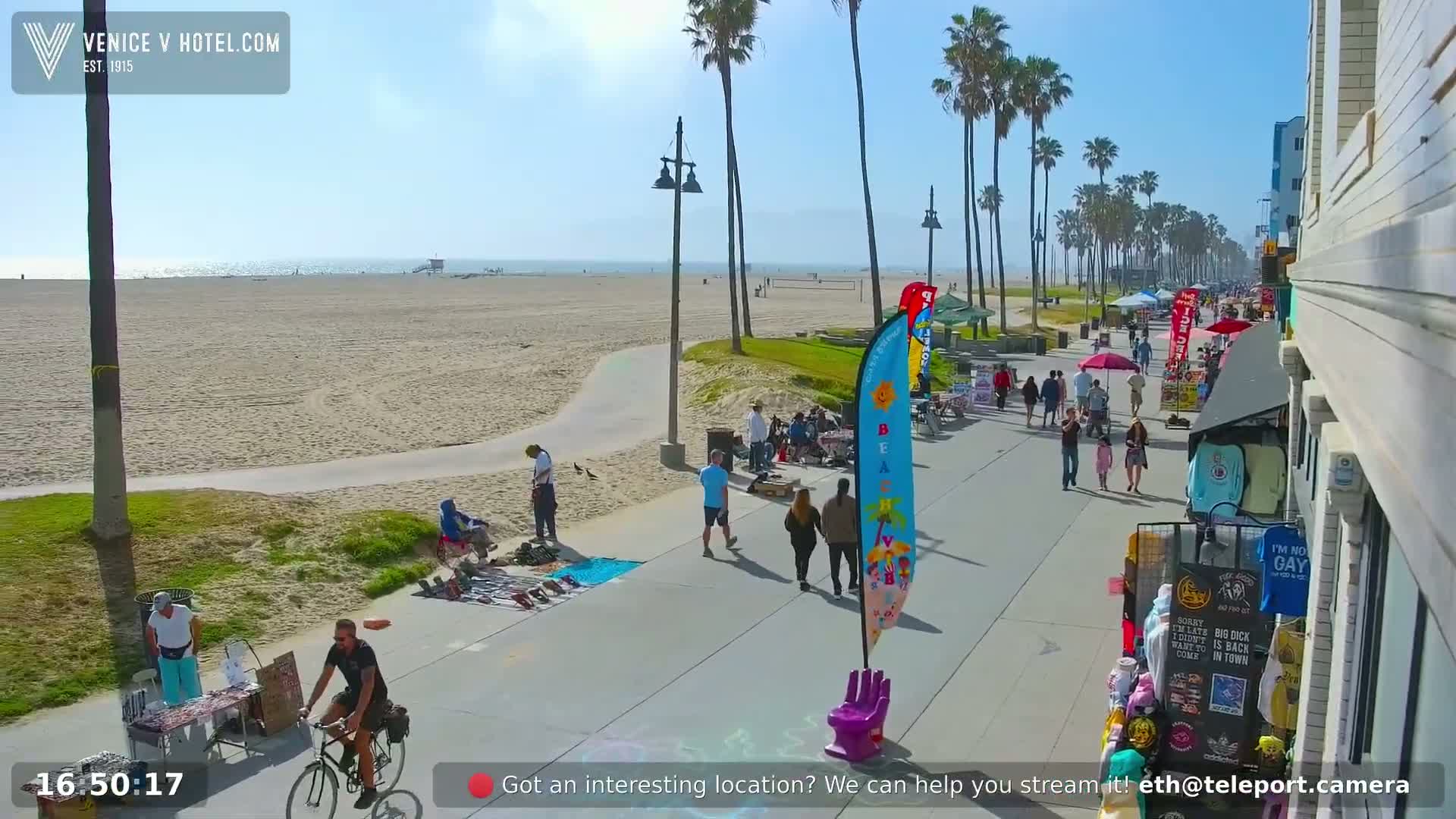 A sunny beach scene shows a paved boardwalk with vendors and pedestrians, palm trees lining the path, and a sandy beach with the ocean in the background.