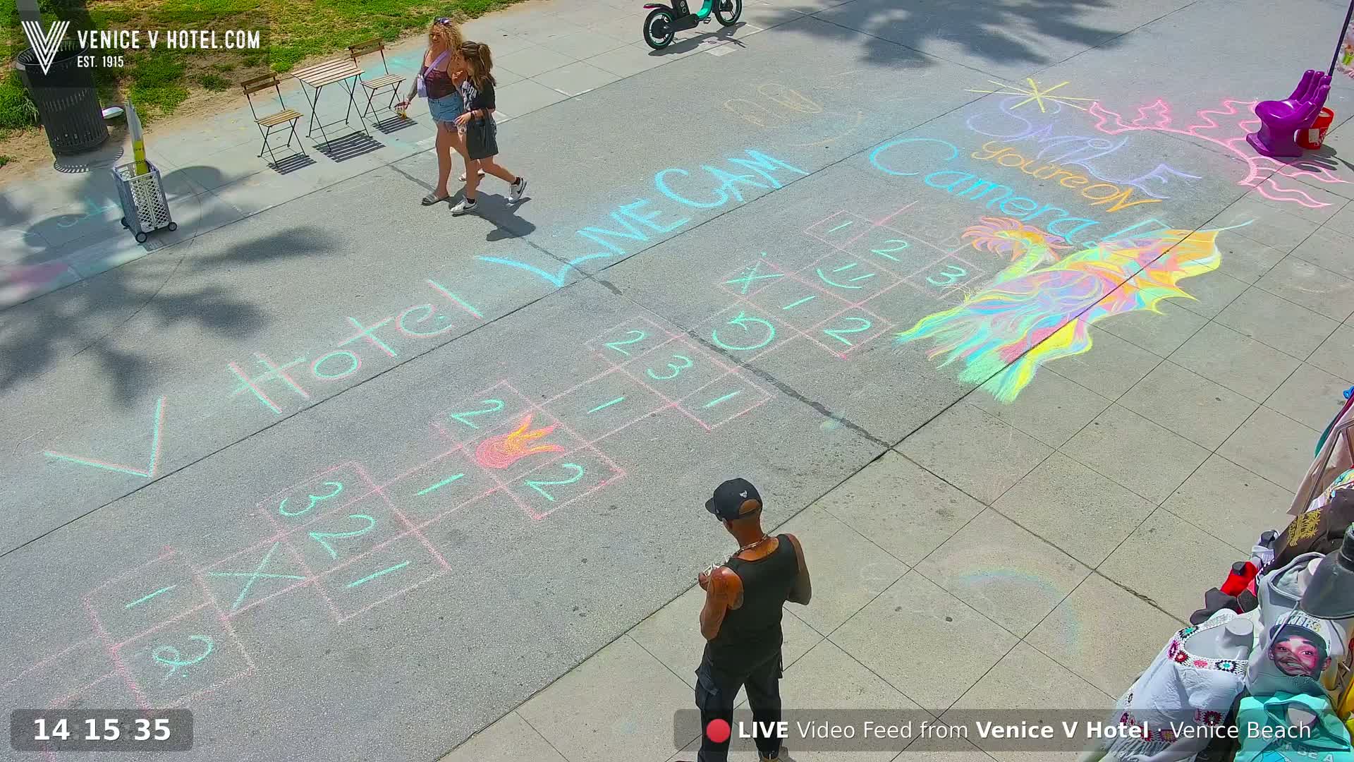Colorful chalk drawings, including games and art, are on a paved surface outdoors on a sunny day, with two people walking and a man standing nearby.