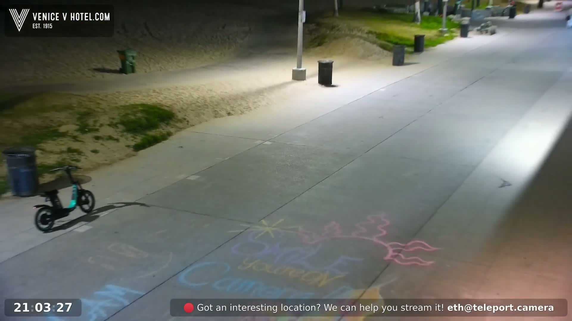 A black electric scooter is parked on a paved path at night, illuminated by streetlights, with chalk drawings on the ground.