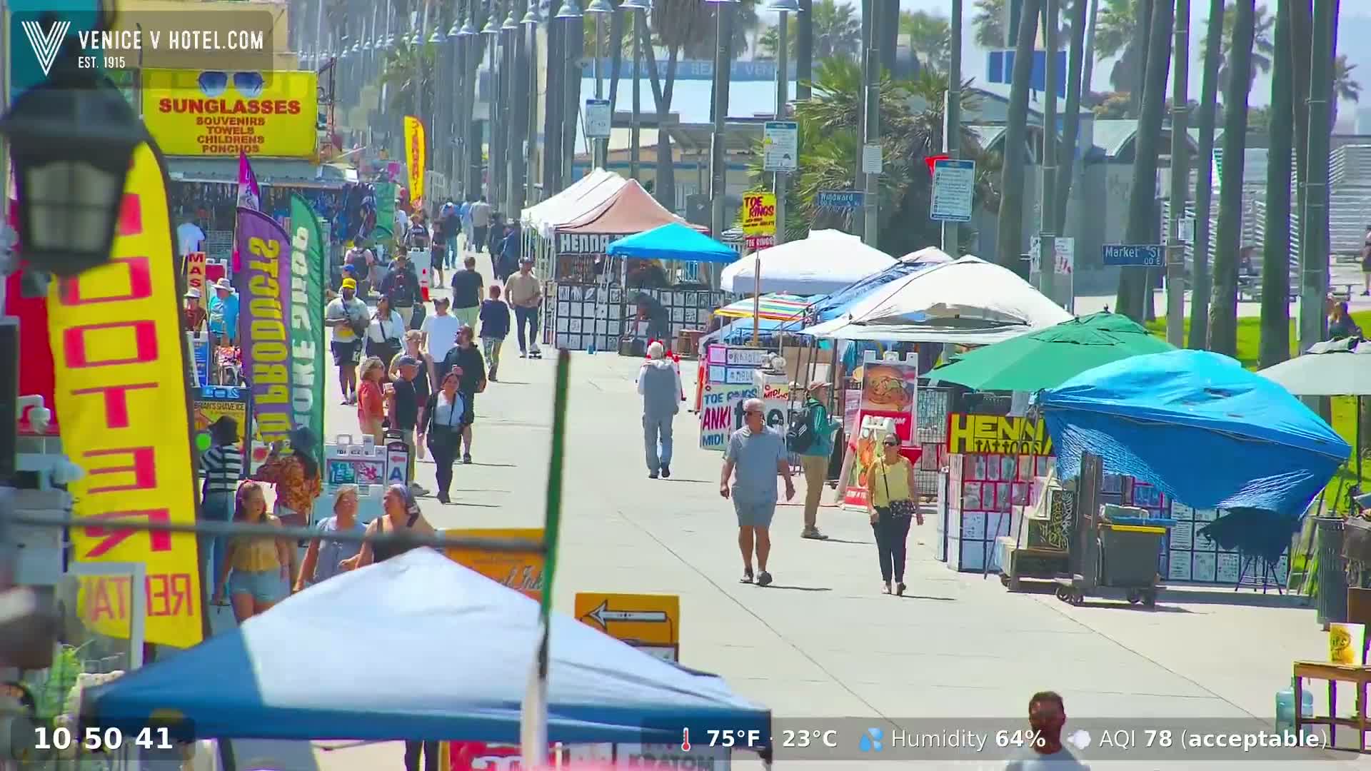 A sunny boardwalk scene shows numerous pedestrians strolling past various vendor stalls under a mostly clear sky.
