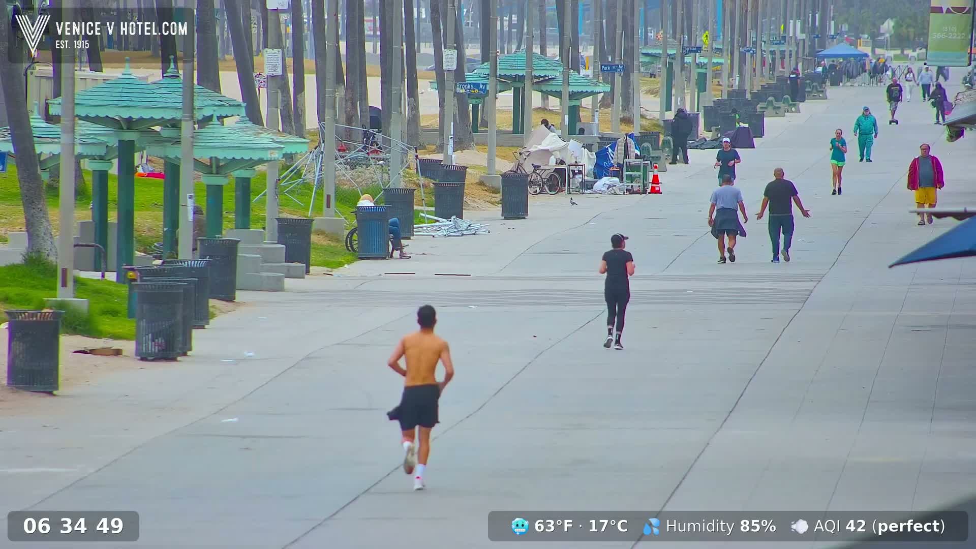 A wide paved walkway lined with palm trees and green park structures shows several people walking and jogging on a slightly overcast day.