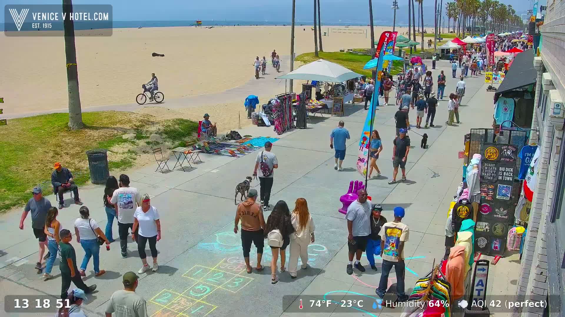 A sunny beach boardwalk scene shows numerous people strolling, shopping at outdoor stalls, and playing hopscotch, with a dog walking nearby and bicycles parked on the sand.