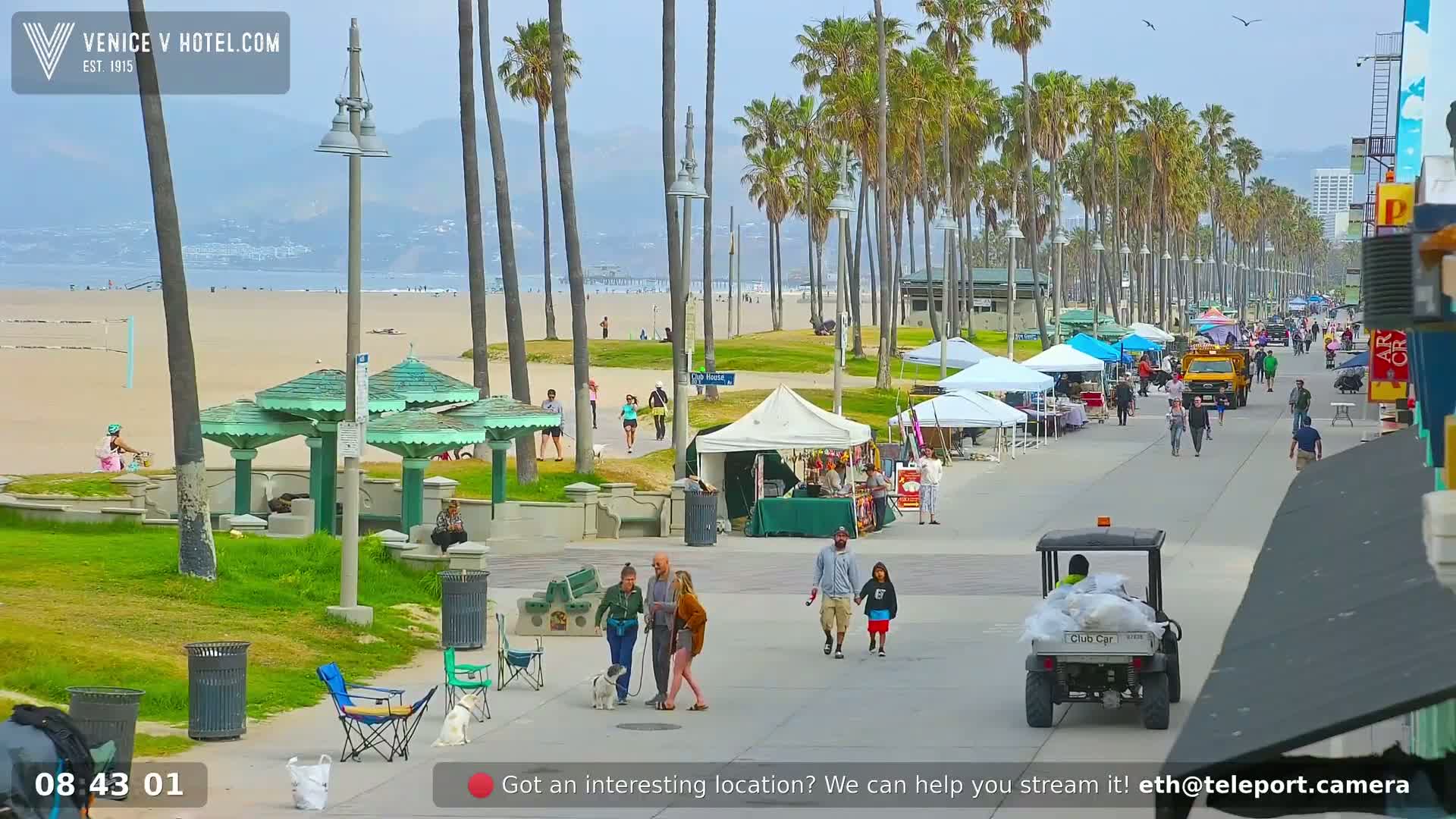 A sunny beach boardwalk scene shows people walking, vendors with tents, and palm trees lining the walkway leading to a sandy beach and ocean in the background.