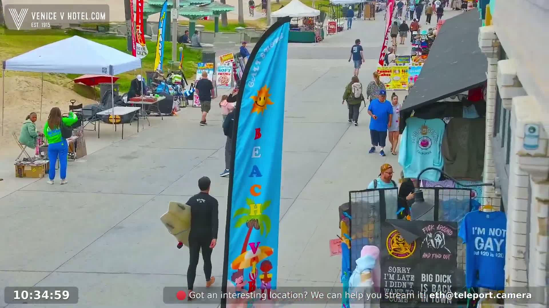 A sunny boardwalk scene shows numerous pedestrians walking past various vendor stalls and beach umbrellas, with a person carrying a surfboard.