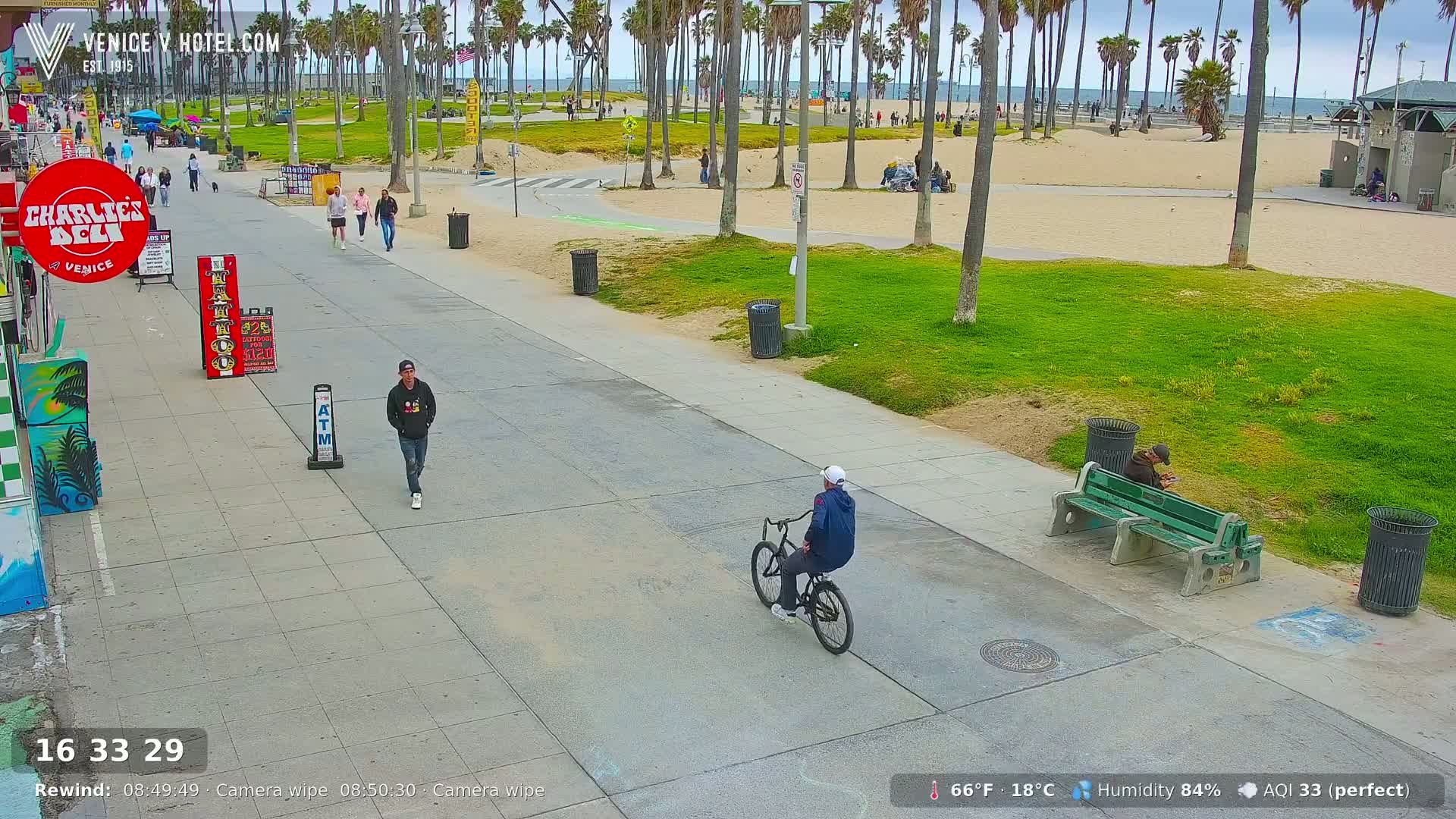 A mostly sunny beach boardwalk scene with palm trees, pedestrians, a bicyclist, and a person sitting on a bench.