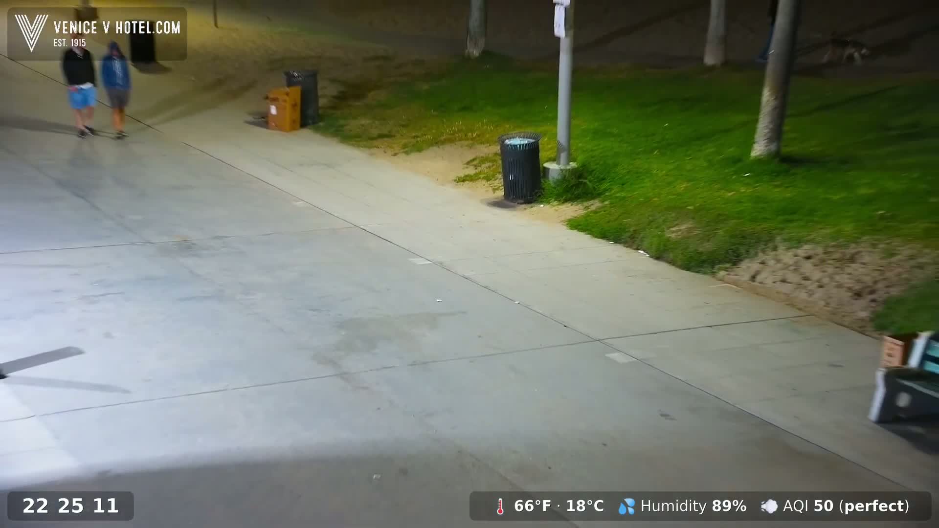 Two people are walking on a sidewalk at night, next to a grassy area with trash cans, under a clear sky with 89% humidity and a temperature of 66°F (18°C).