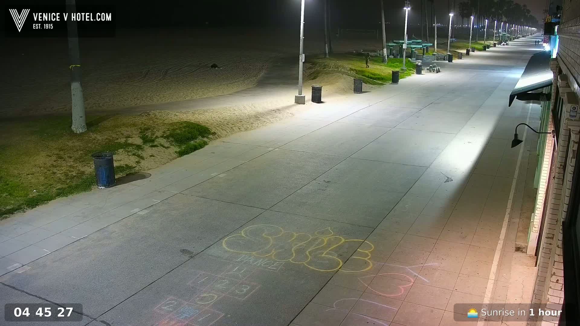 A nighttime view of an empty paved walkway next to a sandy beach, illuminated by streetlights.