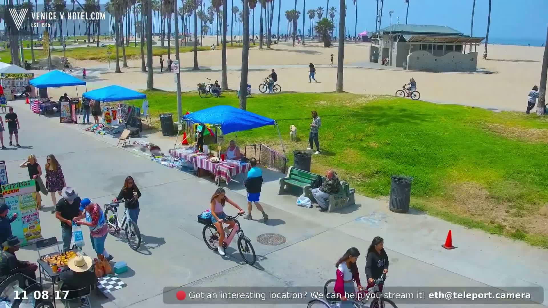 A sunny day at a beach boardwalk shows people walking, bicycling, and vendors selling goods under canopies.