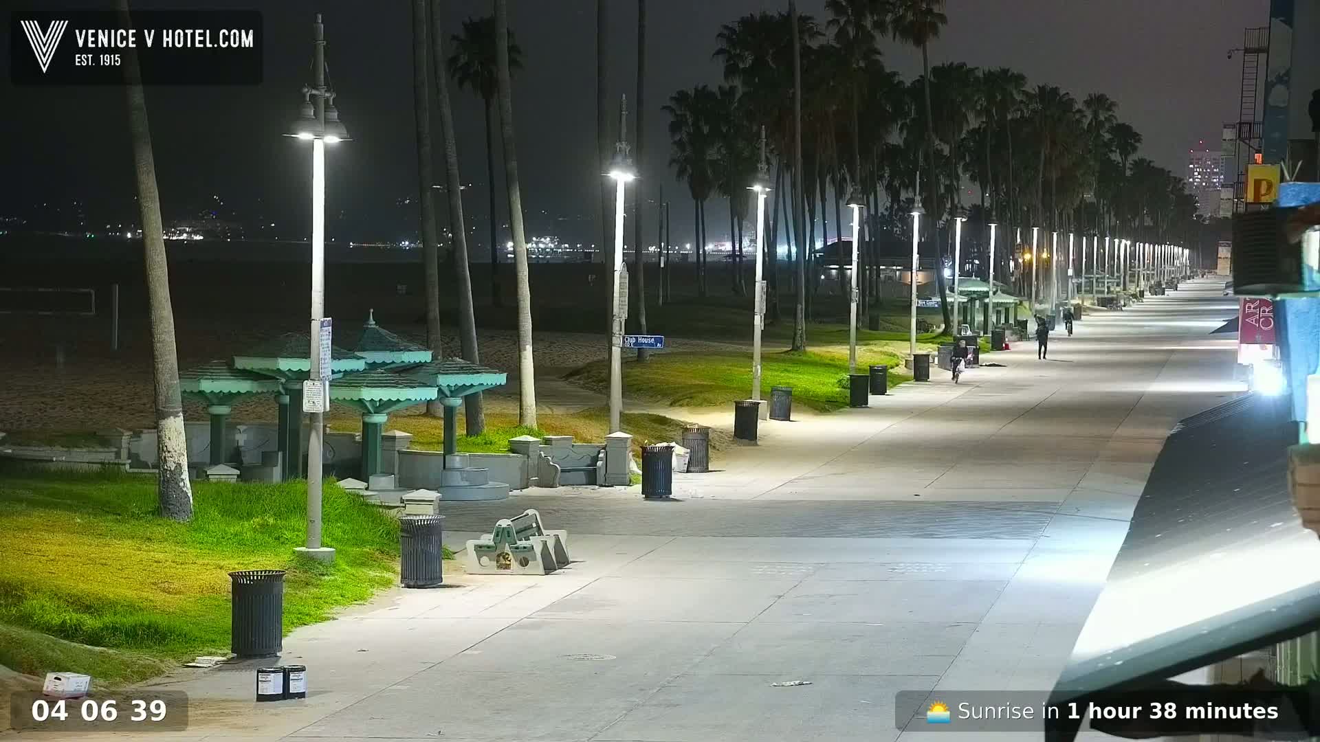A nighttime view of a beach boardwalk lined with palm trees and lights, showing a sandy beach and ocean in the background.