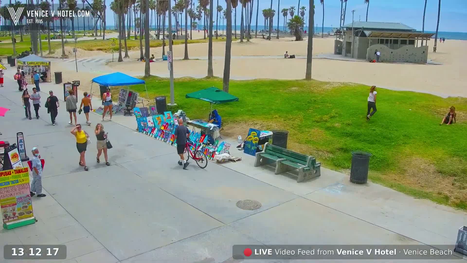 A sunny day at Venice Beach shows pedestrians walking along a boardwalk near vendors and artists, with a beach and ocean visible in the background.