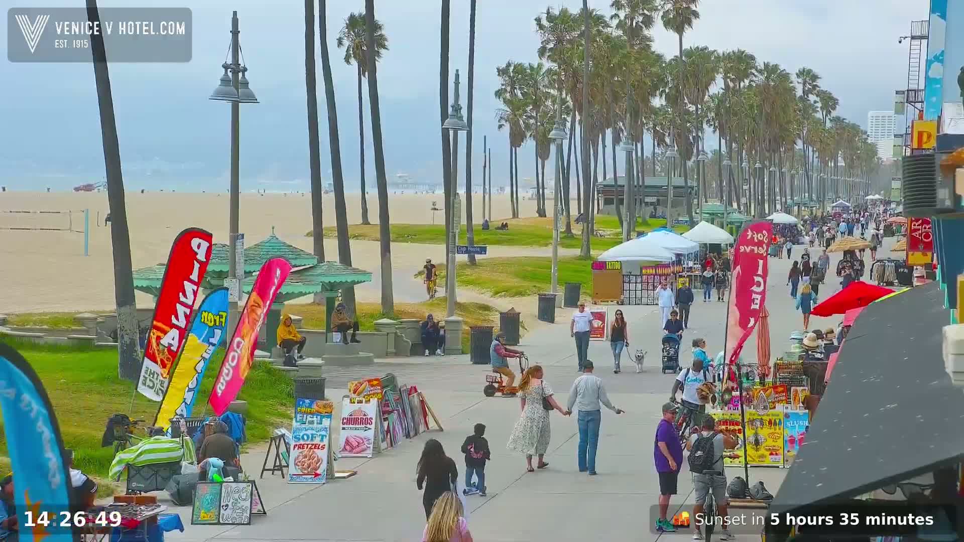A beach boardwalk scene on an overcast day, shows many people walking along a path lined with palm trees, food vendors, and beachgoers enjoying the sand and ocean in the distance.
