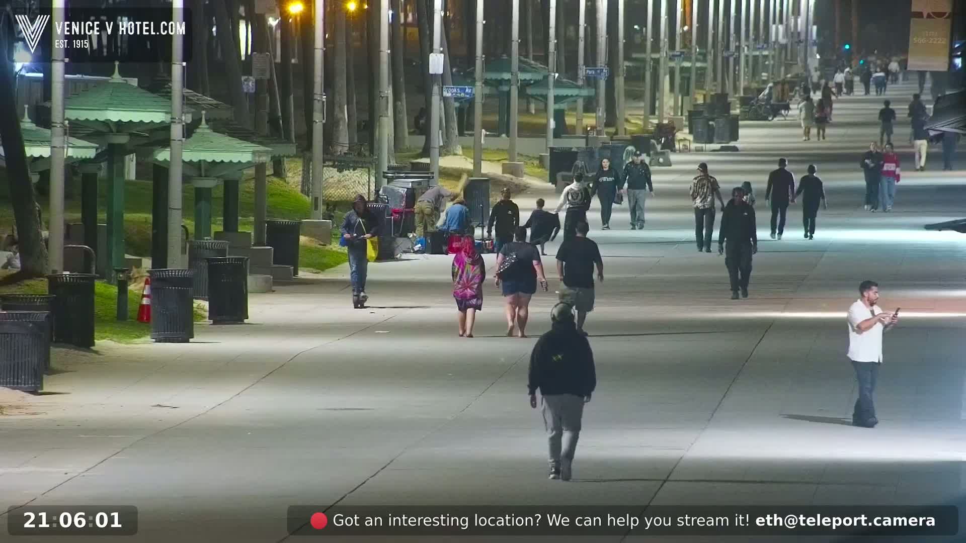 A paved walkway at night, lined with trees and light posts, shows several people walking in different directions under well-lit conditions.