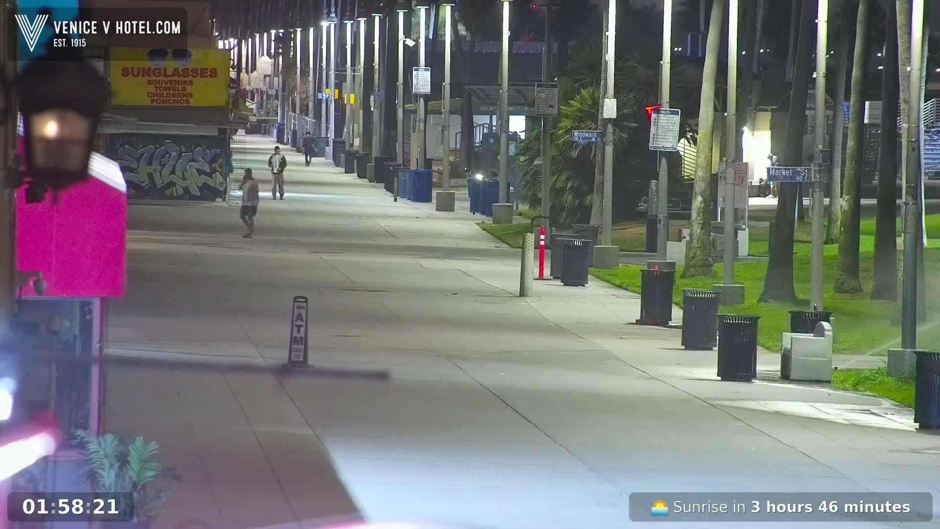 A mostly empty nighttime boardwalk, illuminated by streetlights, with a few pedestrians and palm trees lining a grassy area.