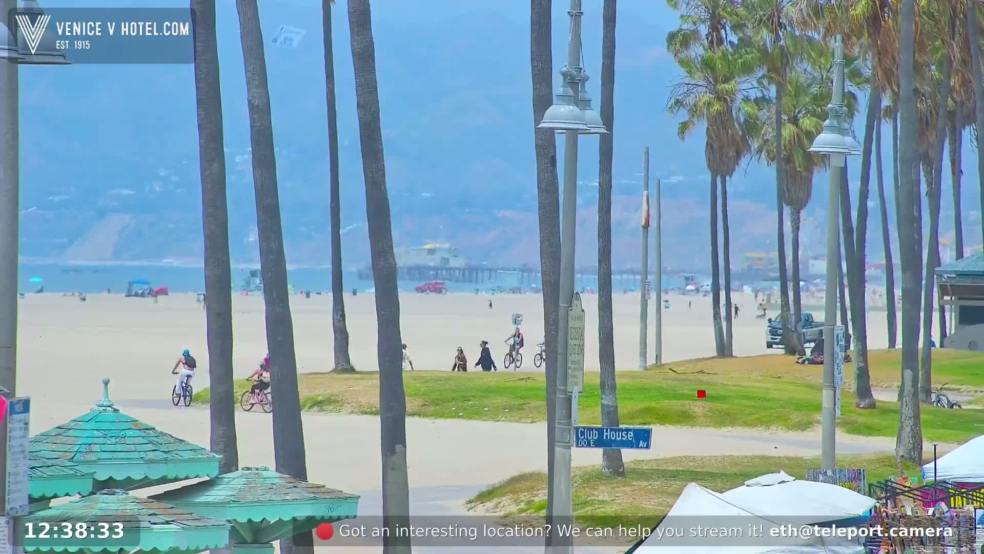 A sunny beach scene shows people walking and biking along a sandy shore lined with palm trees, a pier in the distance, and a small grassy area.