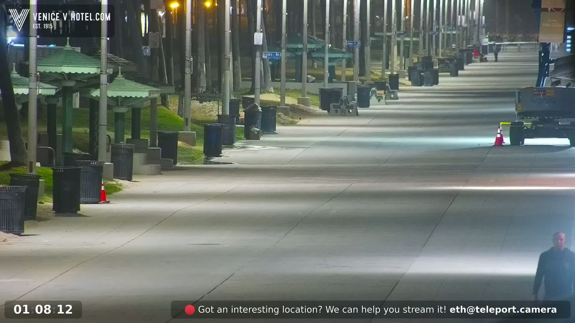 A long, empty paved walkway at night, lined with trash cans and park benches under lampposts, stretches toward a person walking in the distance.