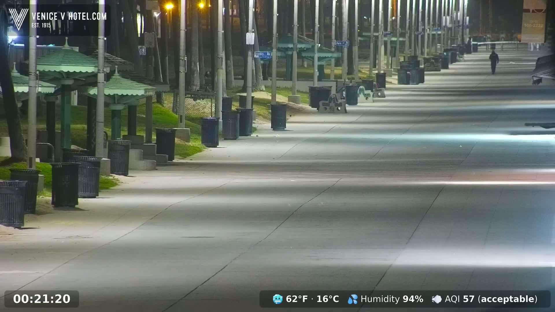 A nearly empty boardwalk at night, illuminated by streetlights, with trash cans and park benches lining the sides, under conditions of 62°F (16°C) and 94% humidity.