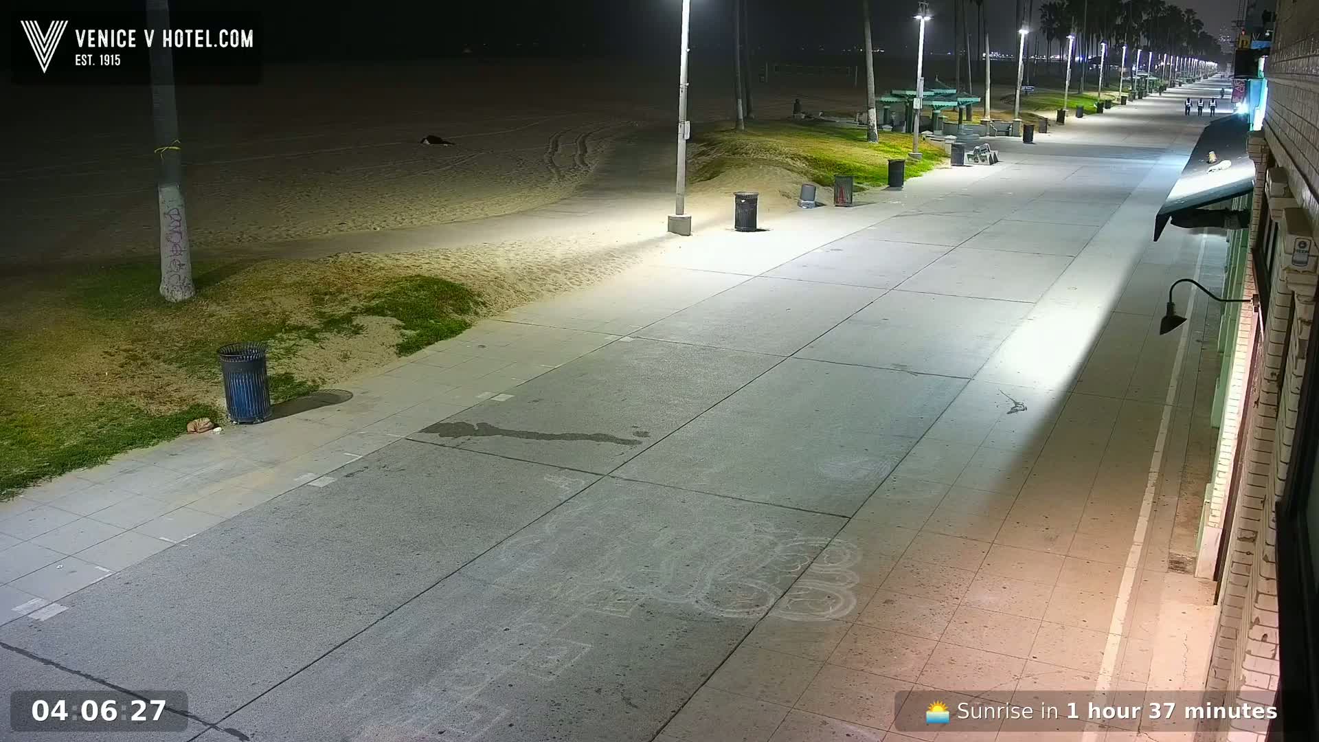 A mostly empty nighttime boardwalk stretches alongside a sandy beach, illuminated by streetlights.