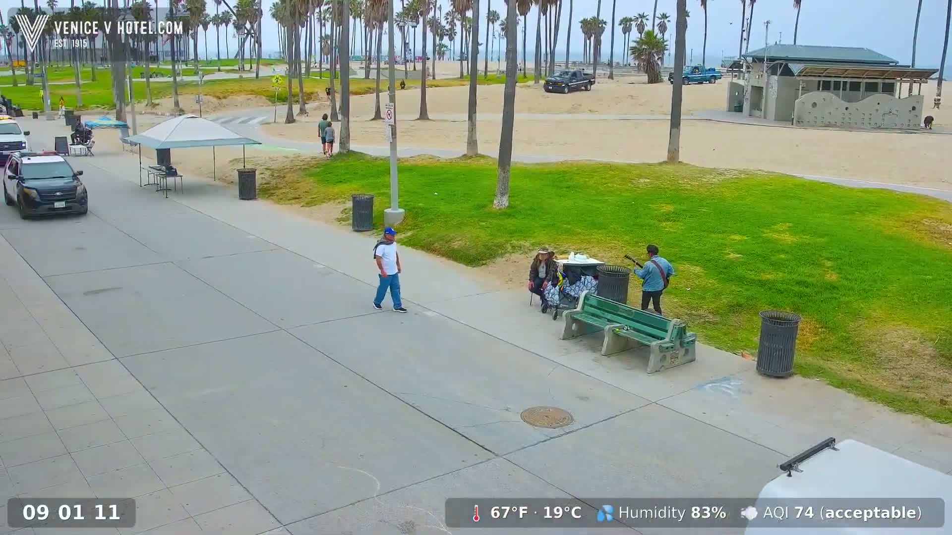 A paved walkway near a beach shows a man walking past a musician playing guitar for a seated group of people under palm trees on a partly sunny day with 83% humidity.