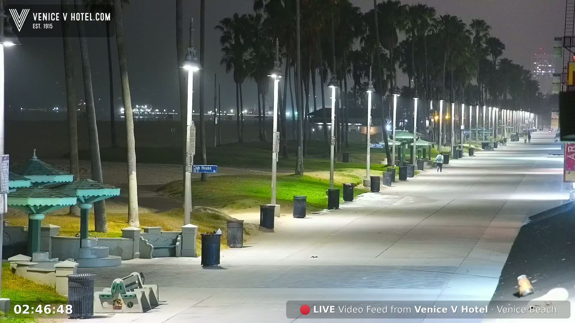 A mostly empty nighttime boardwalk lined with palm trees, lampposts, and trash cans stretches toward a dimly lit beach under hazy conditions.