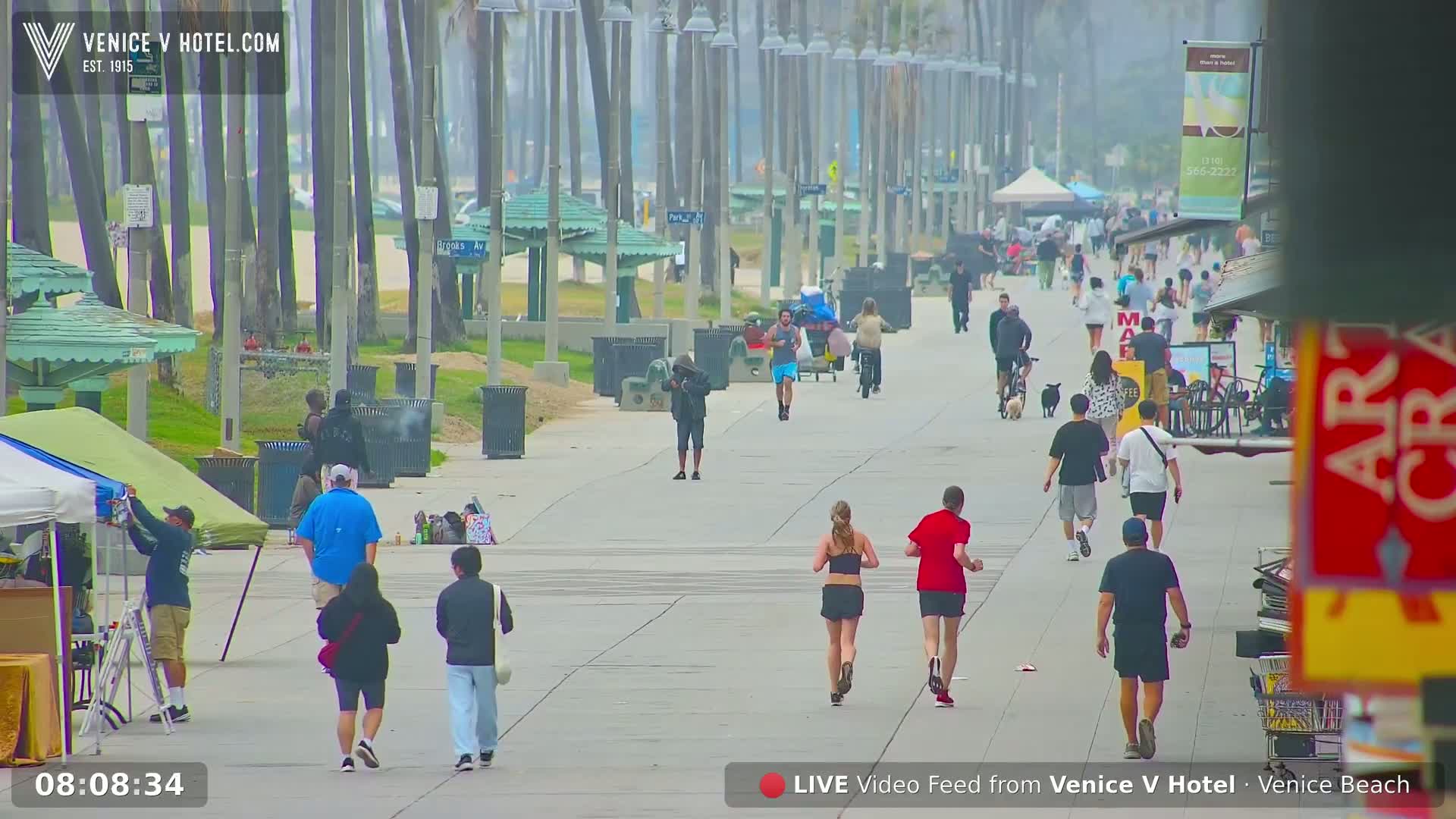 A hazy, sunny day on a beach boardwalk shows numerous people walking, jogging, and biking, with some vendors and beachside shops visible.