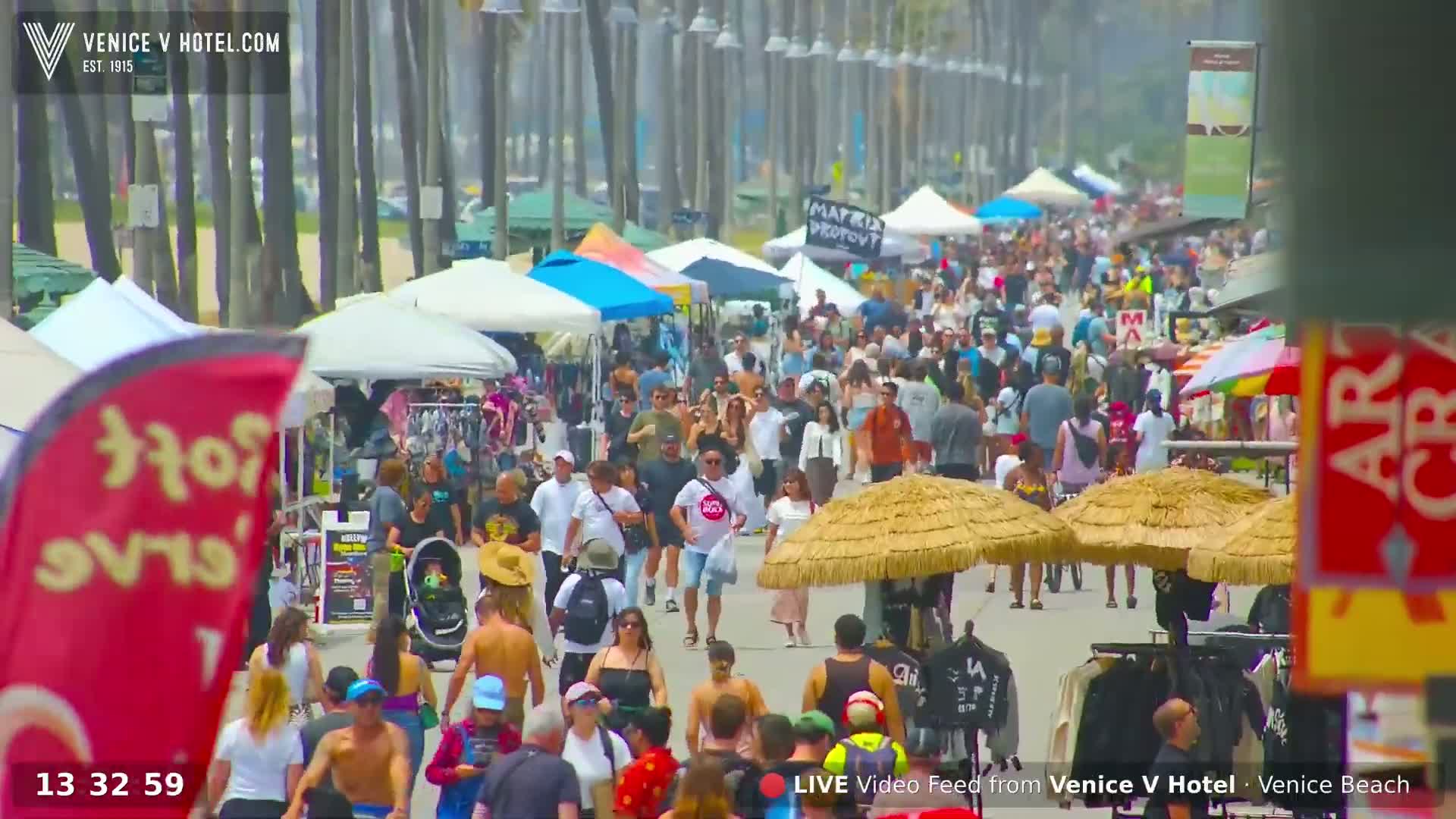 A hazy, sunny day on a beach boardwalk shows numerous people walking, jogging, and biking, with some vendors and beachside shops visible.