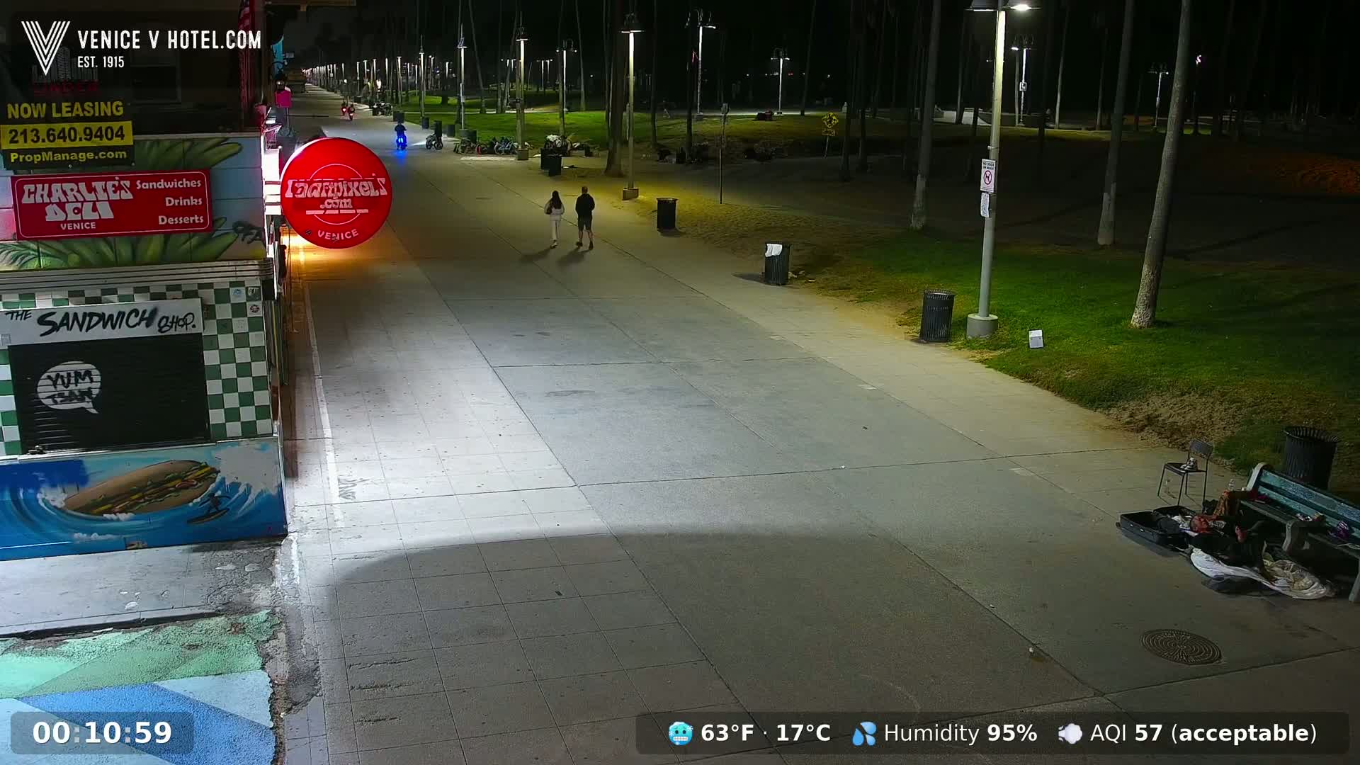 A nighttime view of a mostly empty boardwalk, with a couple walking and a person sleeping on a bench next to a grassy area under the light of street lamps; the humidity is 95%.