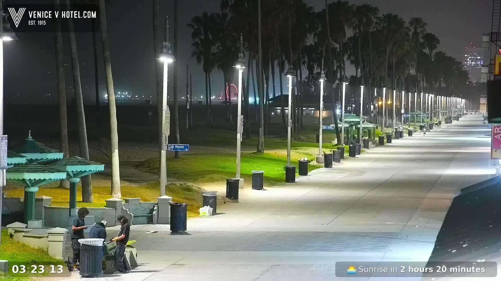 A nighttime view of a beach boardwalk lined with palm trees, lights, and trash cans, with a few people near covered seating areas in the foreground.