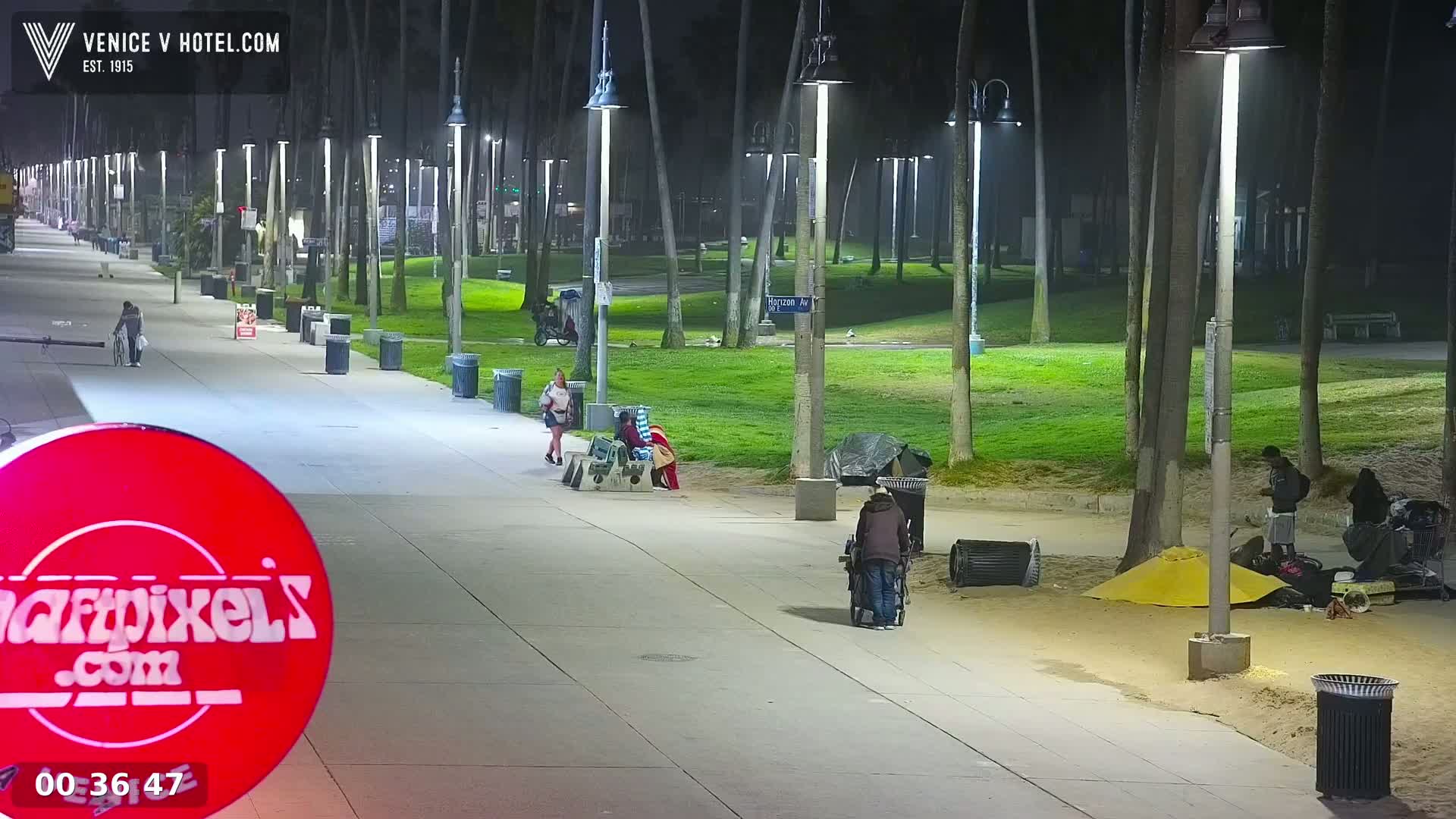 A paved walkway at night, lined with palm trees and streetlights, shows several people walking and some people sitting near makeshift shelters on the sandy ground beside it.
