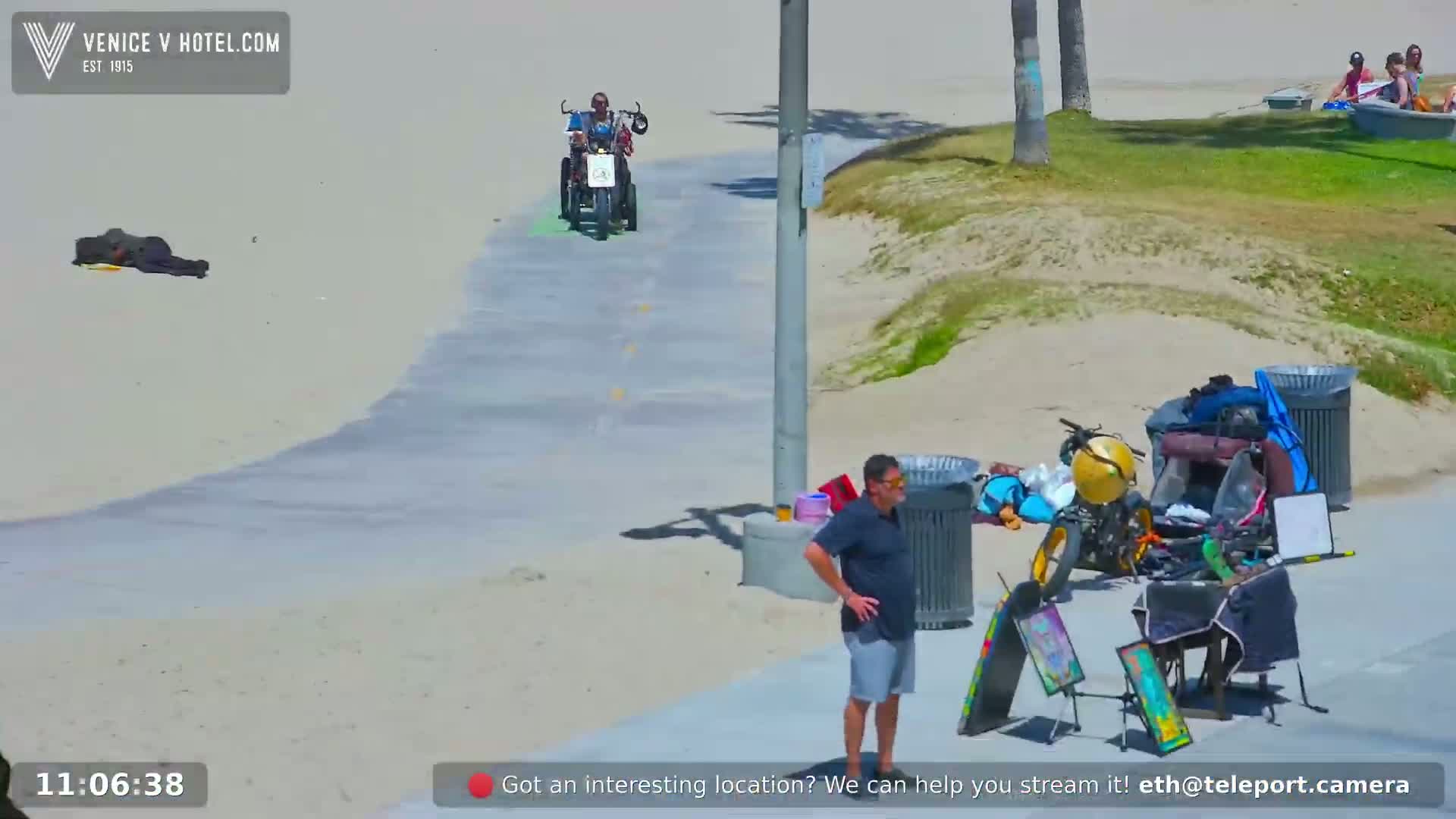 A person on a three-wheeled vehicle rides along a paved path next to a sandy beach on a sunny day, near a man standing and various items by trash cans.