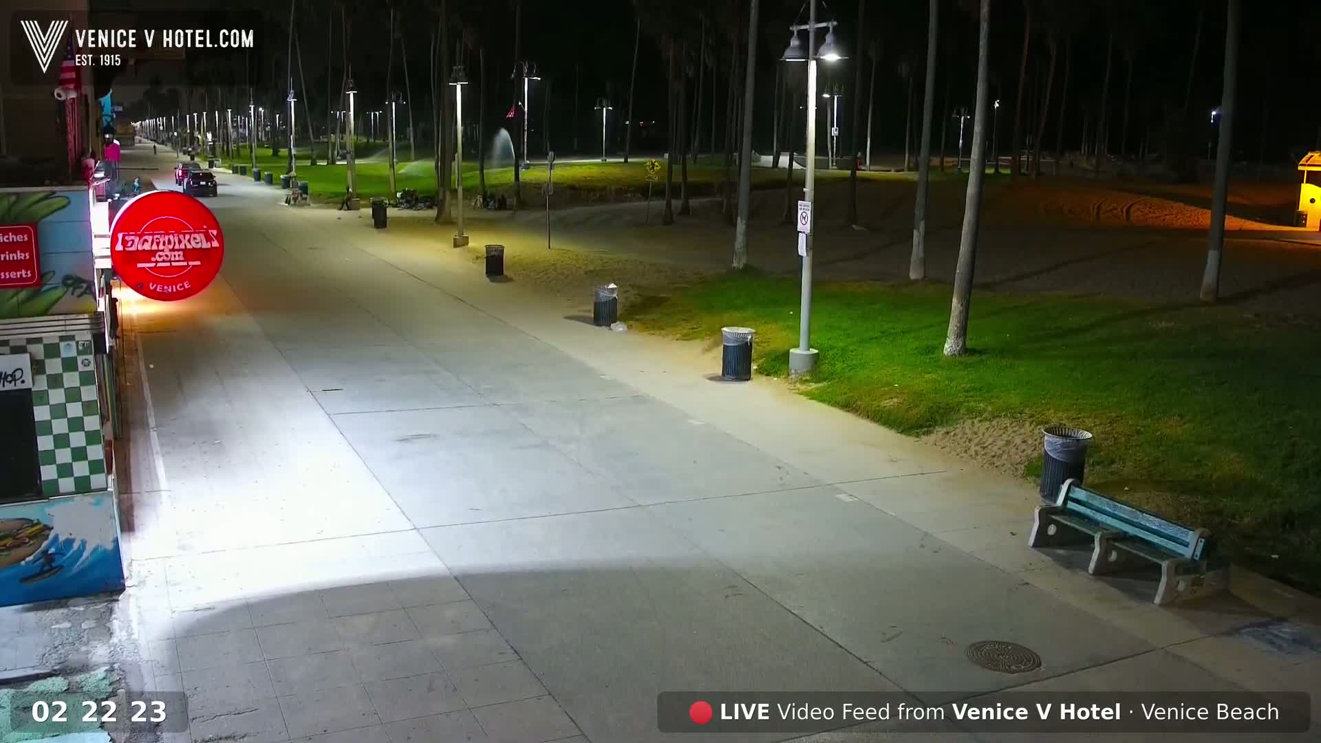 A mostly empty nighttime boardwalk, lined with palm trees and lampposts, stretches toward a beach.