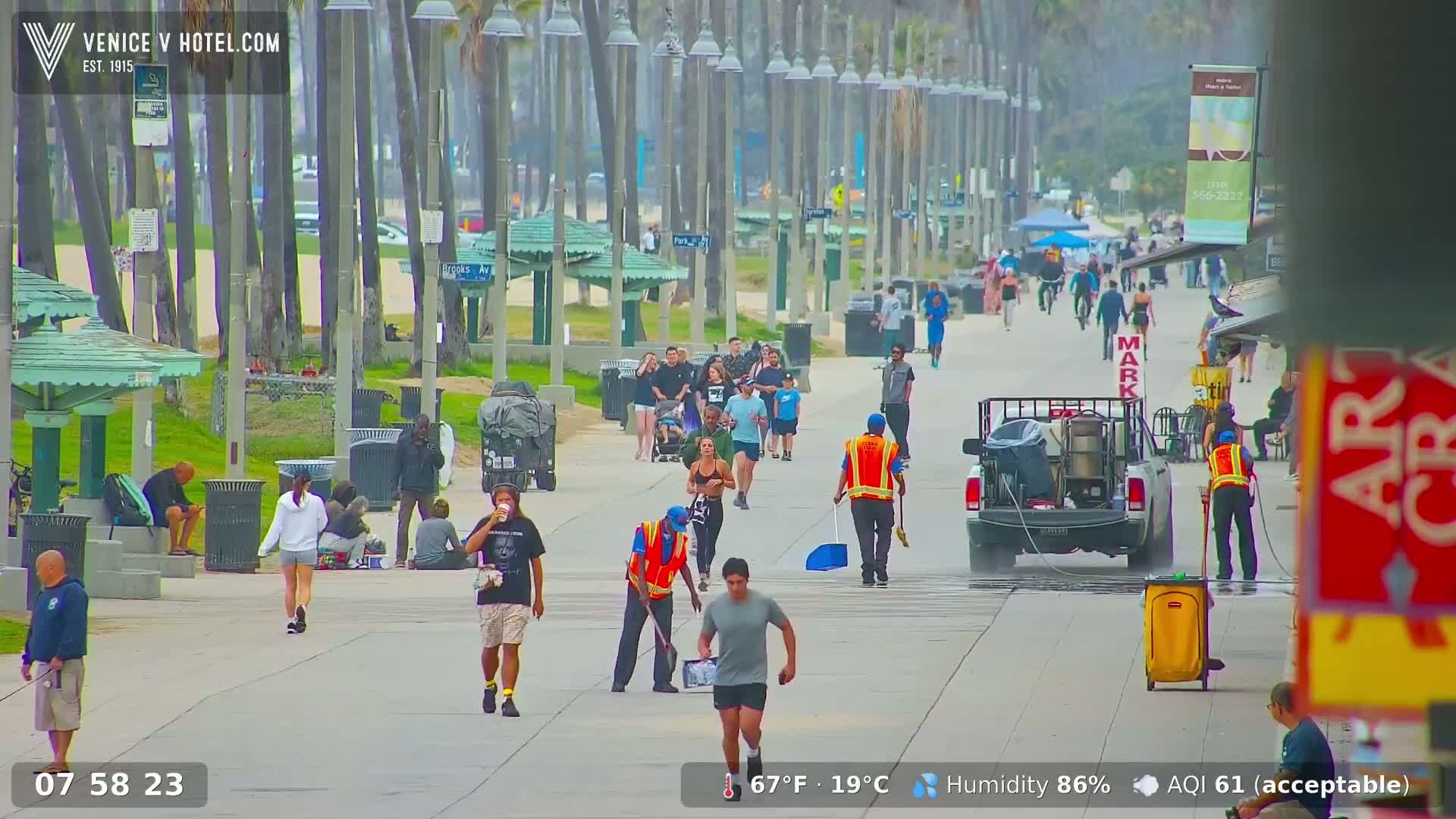 A paved boardwalk, lined with palm trees and beachside amenities, is populated with pedestrians and street cleaners on a humid day.