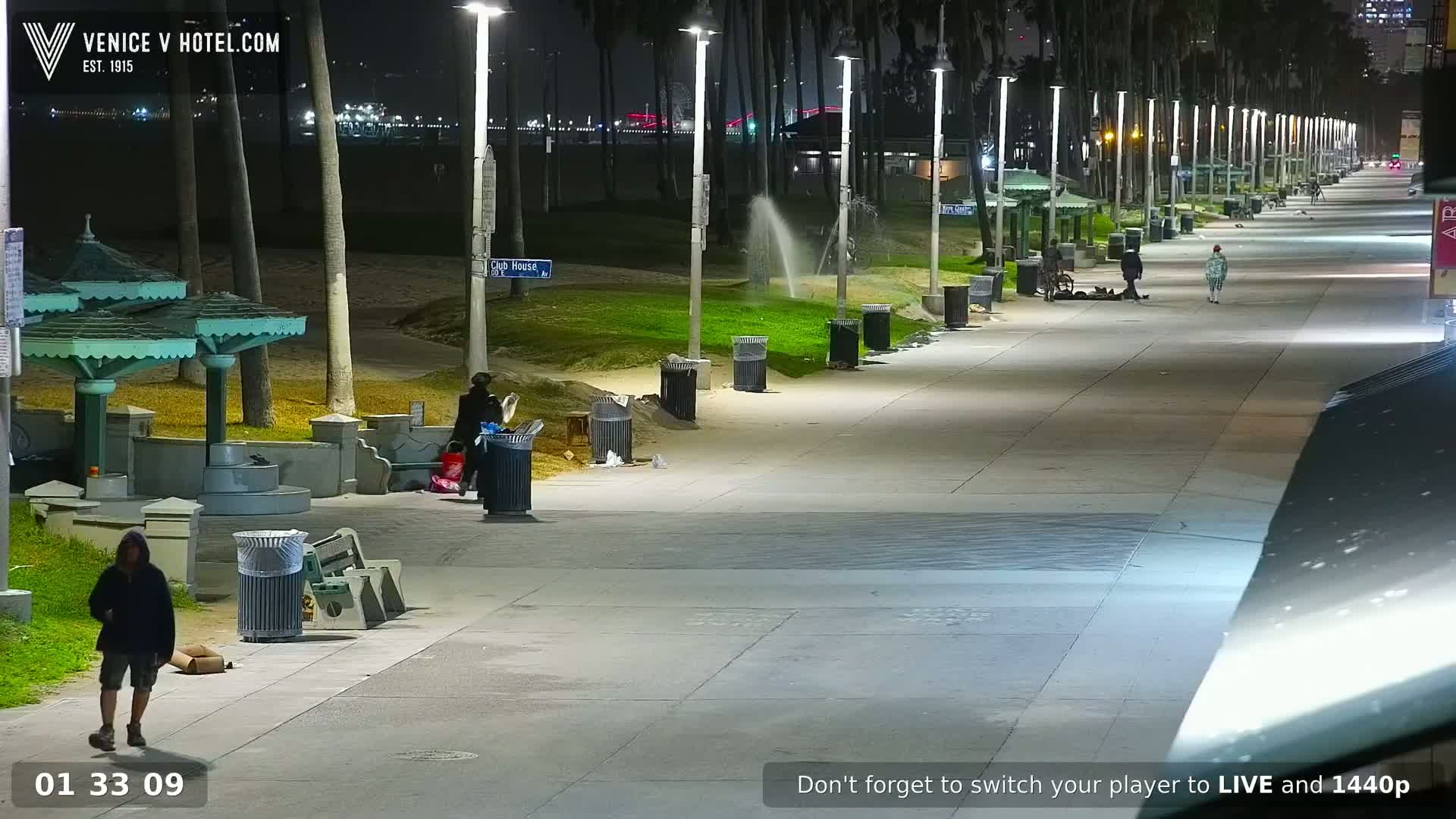 A nighttime scene of a mostly empty boardwalk lined with palm trees, lights, and trash cans, with a few people walking and some sitting.