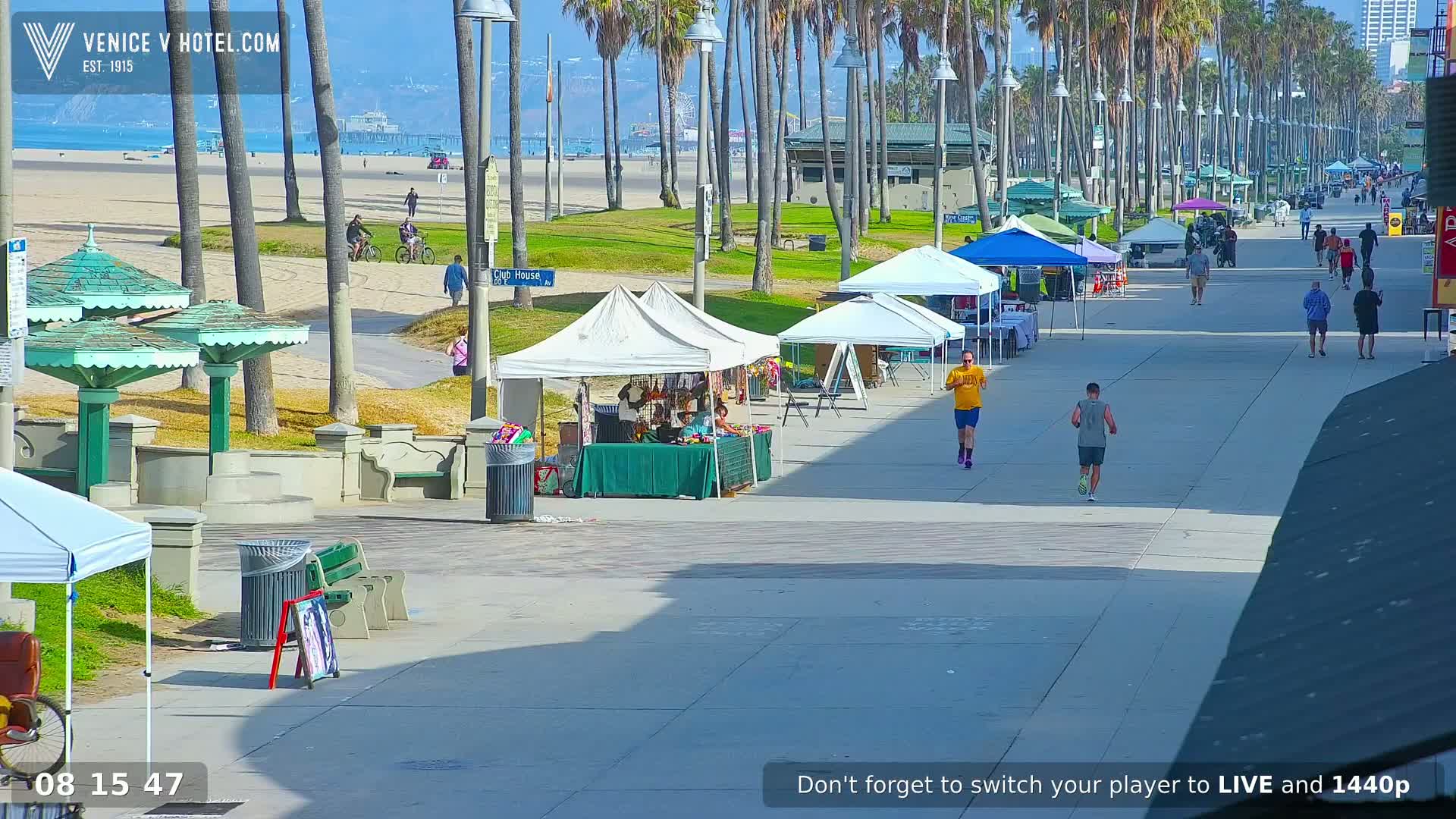 A sunny day at a beach boardwalk shows people walking and jogging past numerous market stalls and beachside amenities under a clear sky.