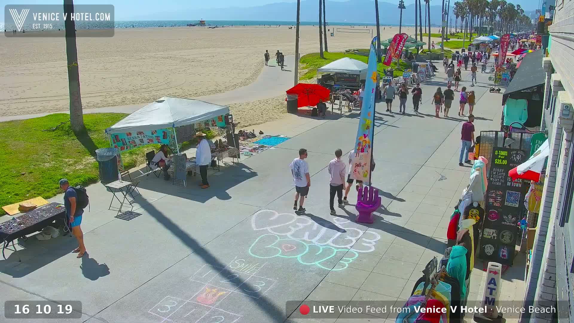 A sunny beach boardwalk scene shows several vendors' booths and people walking past chalk art on the pavement, near a sandy beach and ocean.