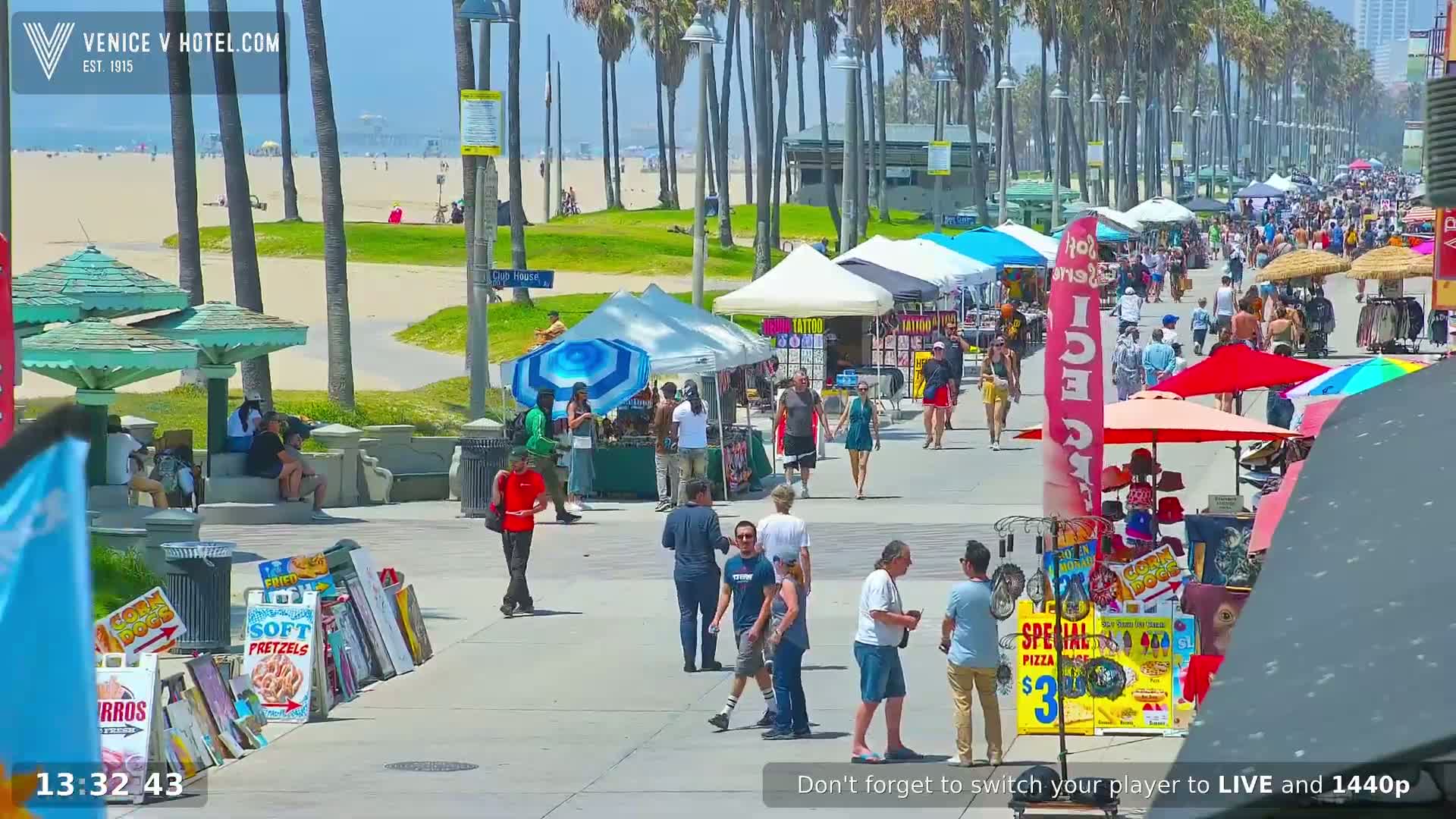 A sunny beach boardwalk scene shows numerous people walking past various vendors and temporary shops under a clear sky.
