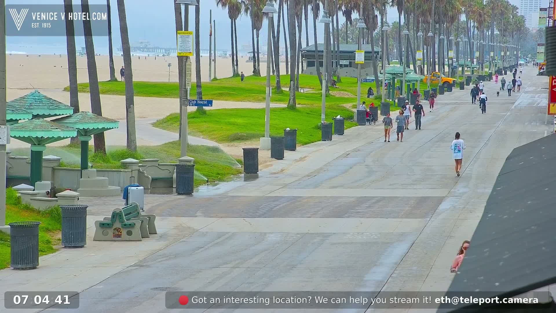 A sunny beach boardwalk scene shows numerous people walking past various vendors and temporary shops under a clear sky.
