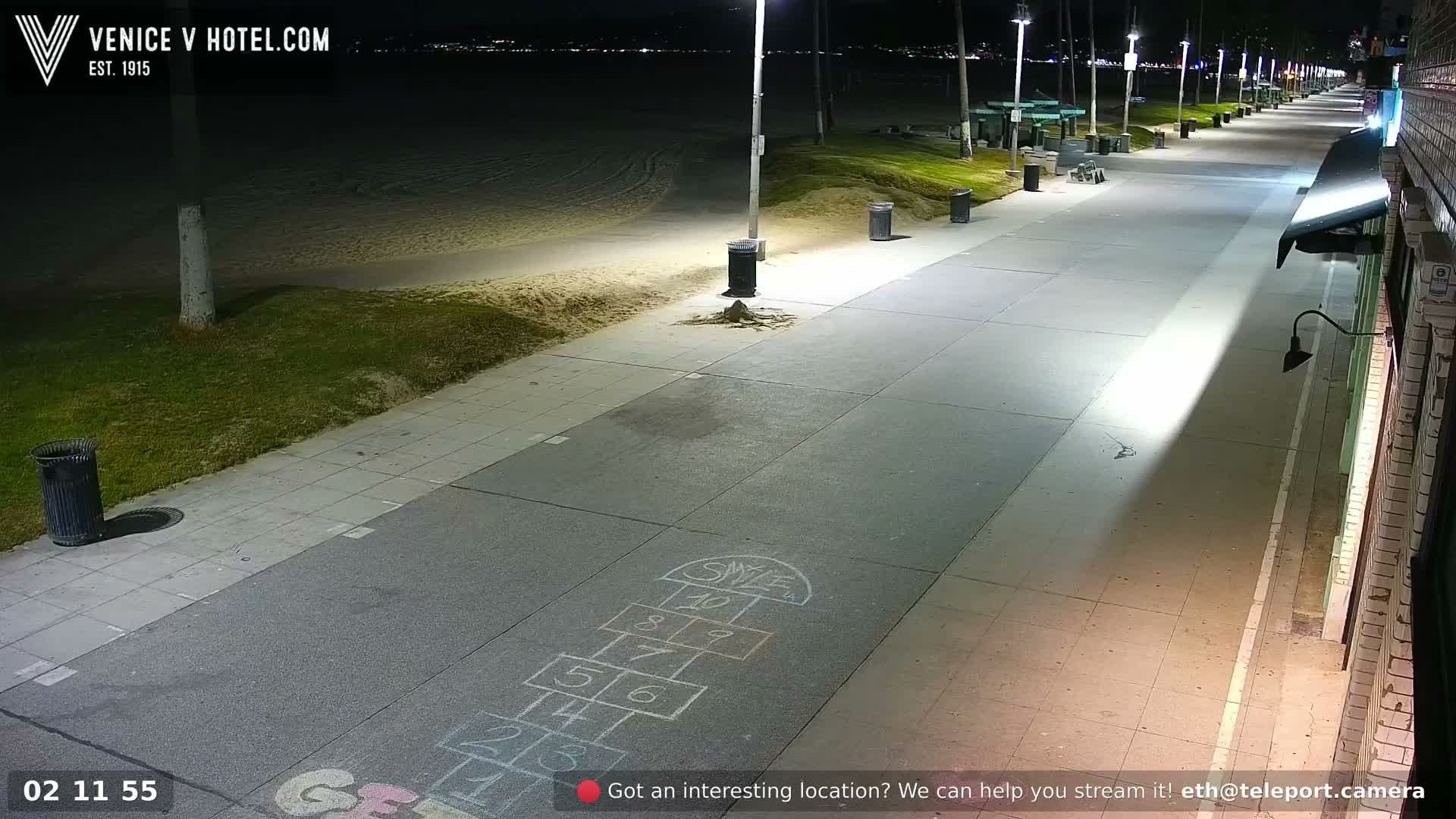 A brightly lit boardwalk with a hopscotch game drawn on the pavement stretches along a beach at night under clear skies, with city lights visible in the distance.