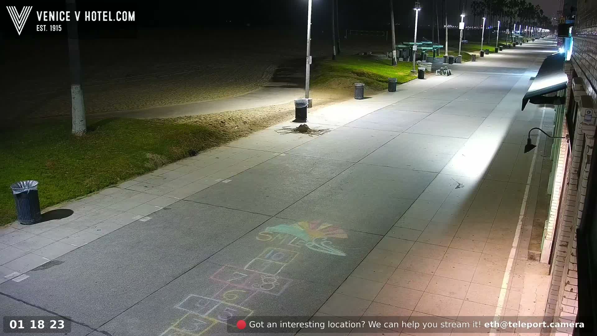 A brightly lit boardwalk with a hopscotch game drawn on the pavement stretches along a beach at night under clear skies, with city lights visible in the distance.