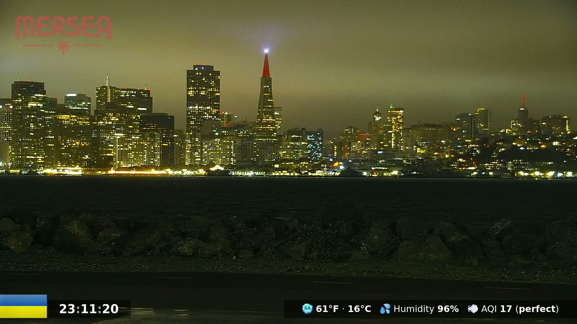 A nighttime view of a brightly lit city skyline across a dark body of water under a hazy sky.