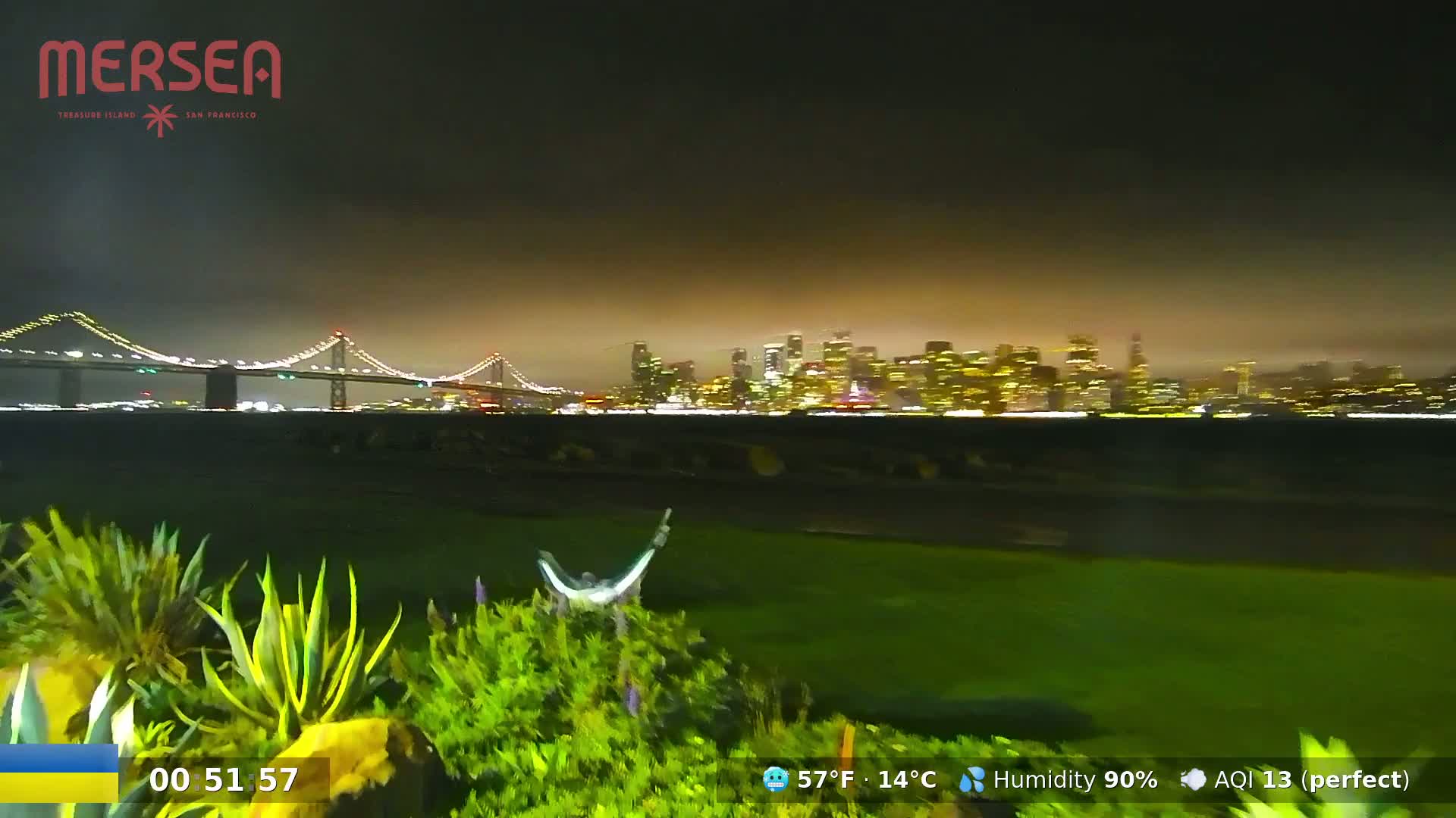 A nighttime view of a lit-up city skyline across a body of water, with a bridge in the foreground under a hazy, humid sky.