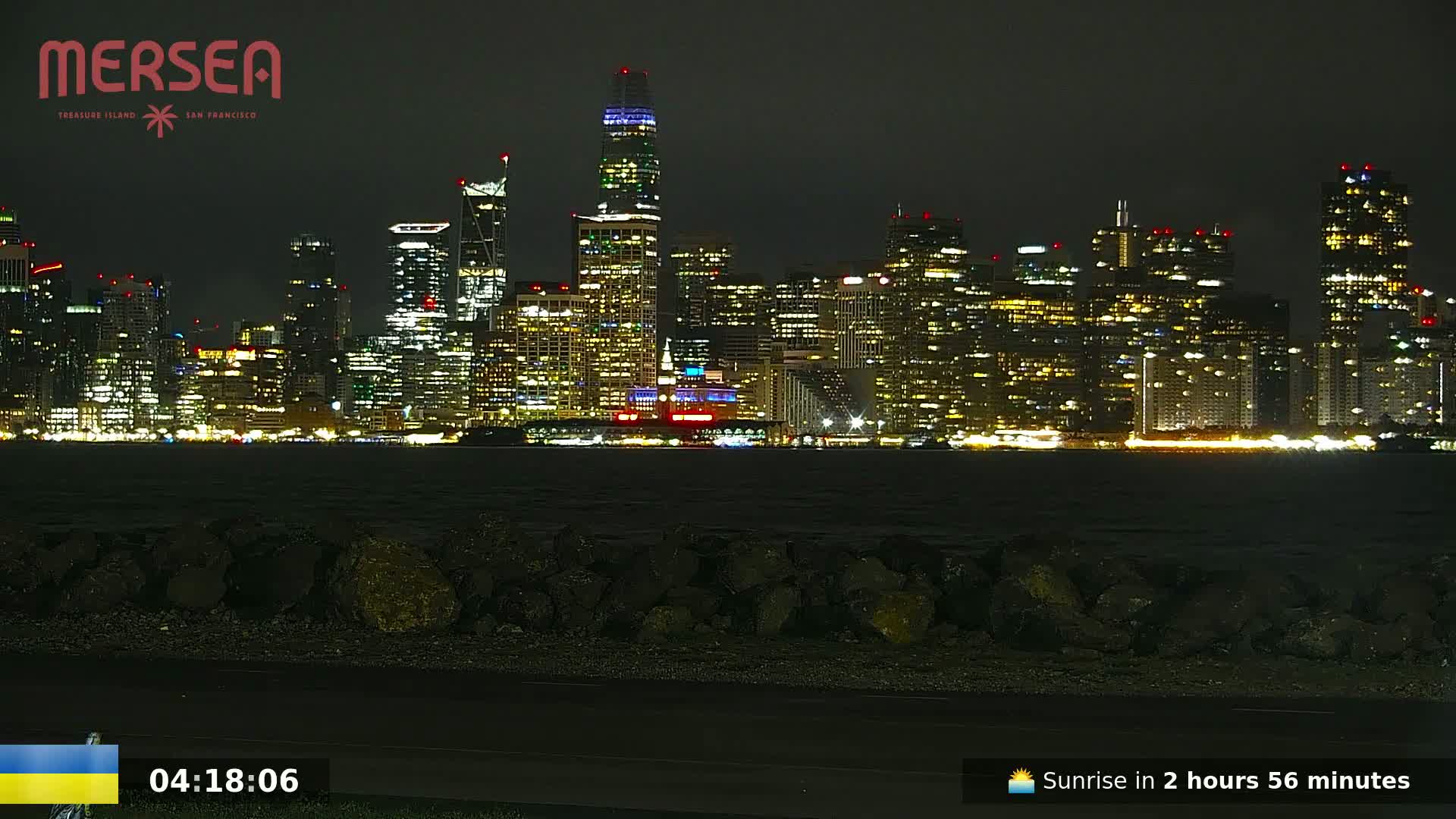 A nighttime view of a city skyline across a dark body of water, with a rocky shoreline in the foreground.