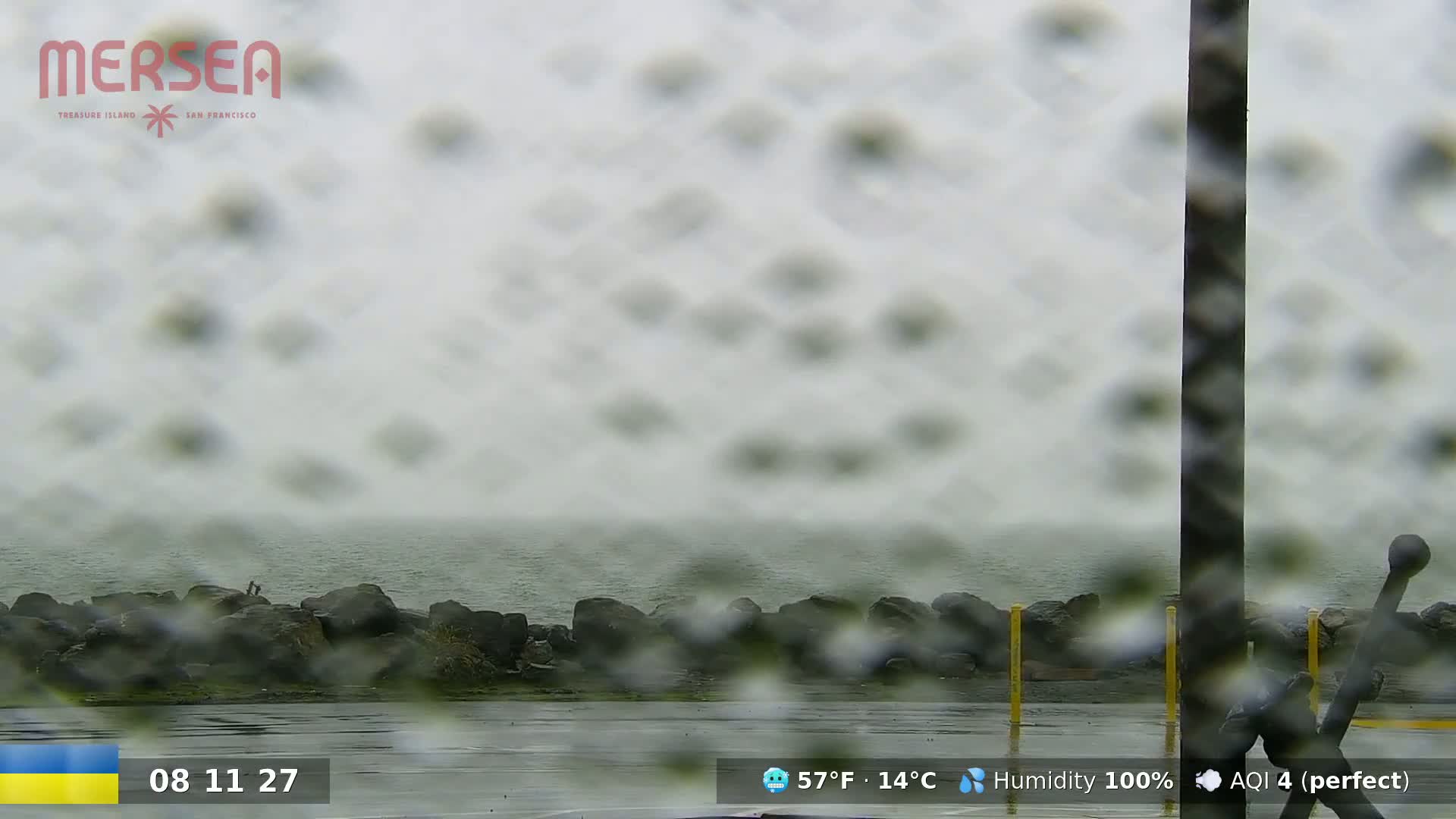 A rain-covered window shows a view of a rocky shoreline and calm water under an overcast sky.