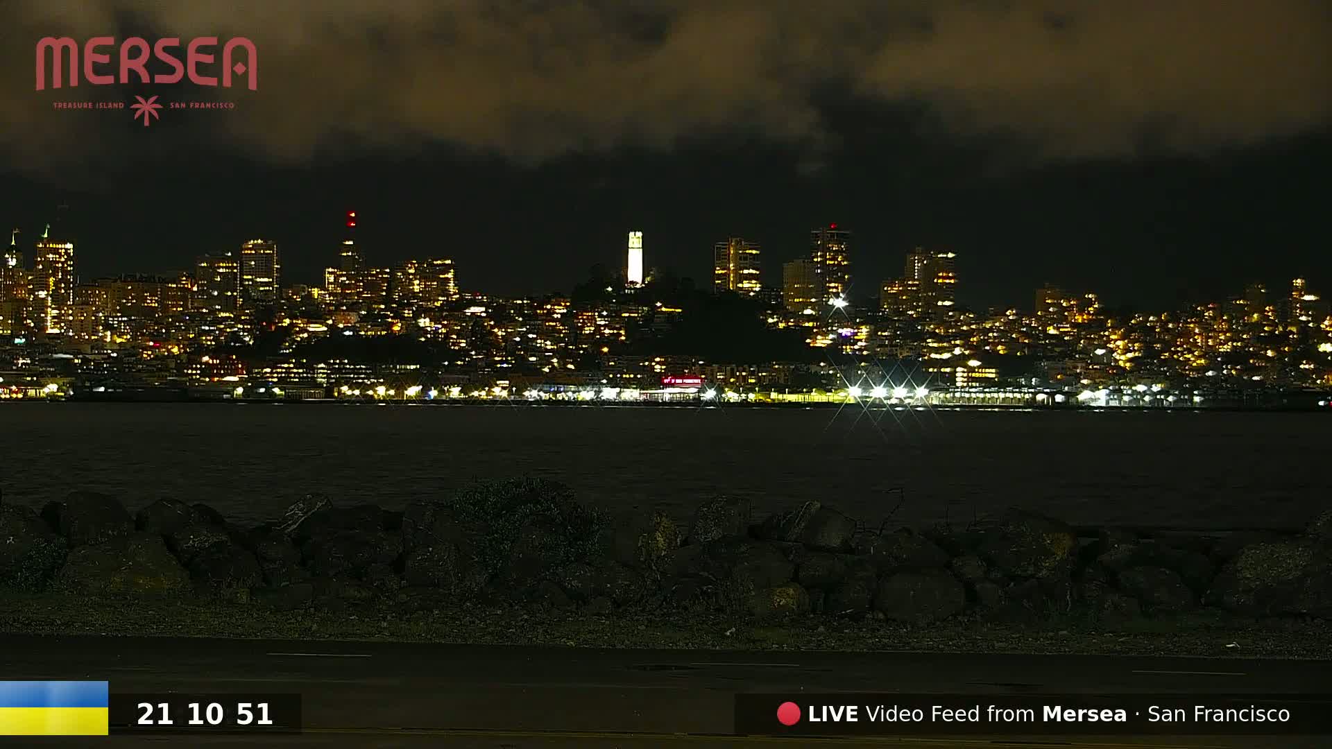 A nighttime view of a city skyline illuminated with lights across a dark body of water under a cloudy sky.