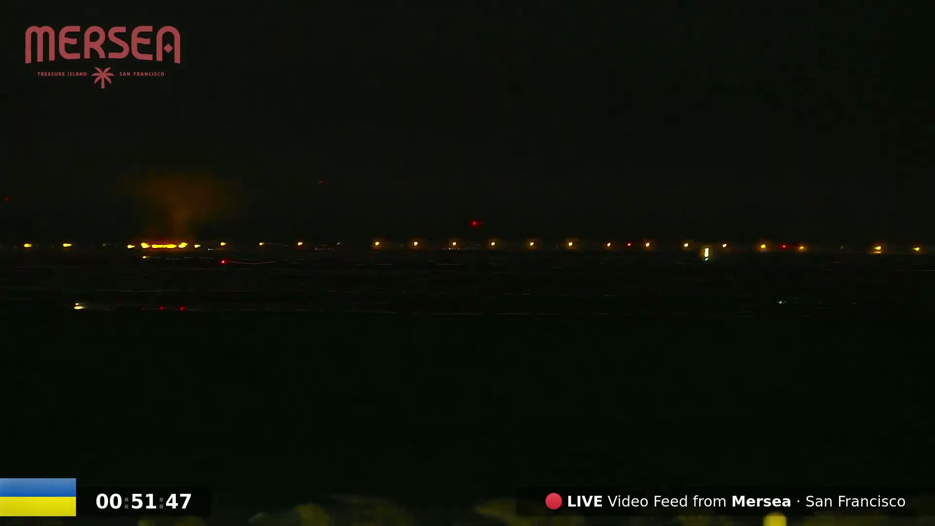At night, a line of lights is visible in the distance across a dark body of water under a dark sky.
