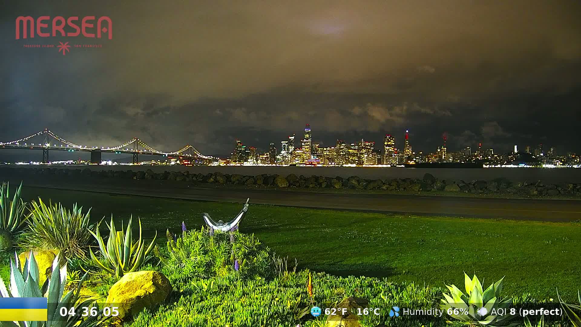 A nighttime view of a city skyline across a body of water, under a cloudy sky, with a bridge in the foreground.