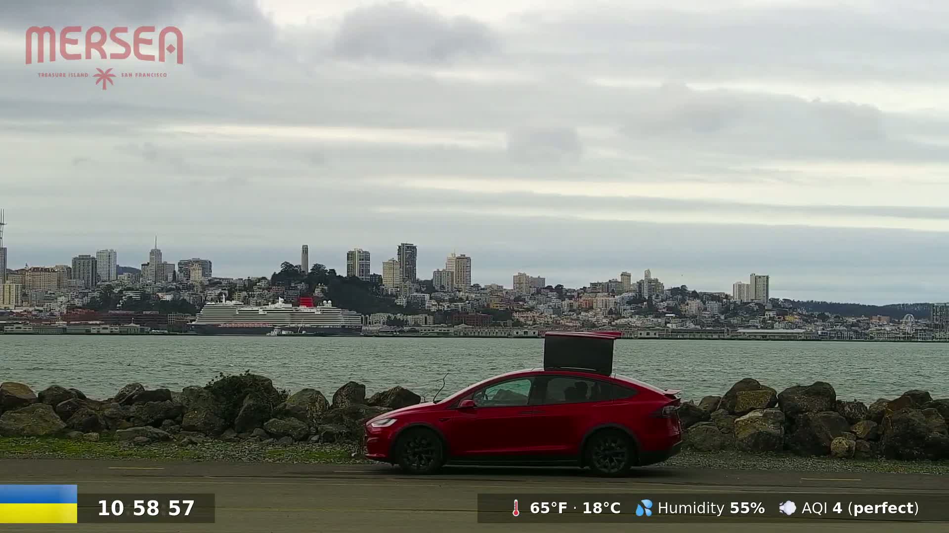 A red Tesla Model X with a rooftop carrier is parked near a rocky shoreline overlooking a city skyline and a cruise ship under a cloudy sky.