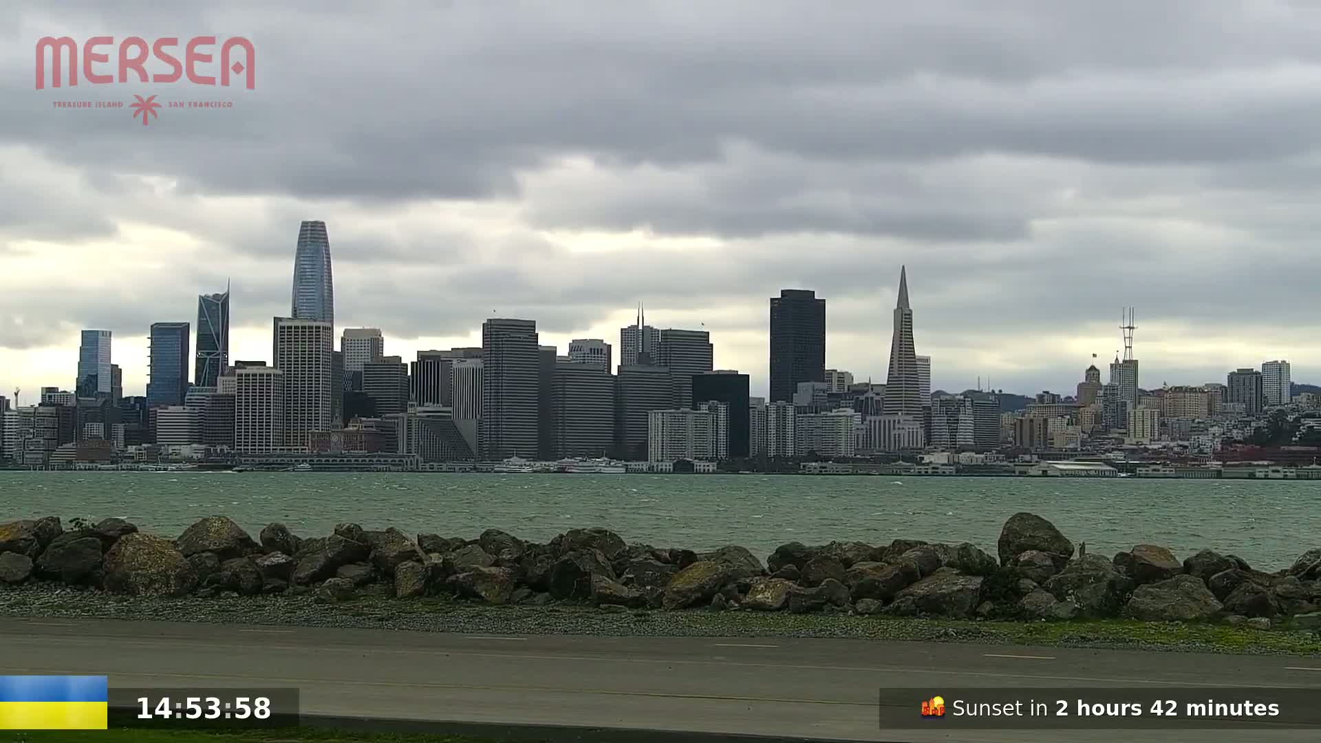 A rocky shoreline and choppy water separates the viewer from a cityscape under a cloudy sky.
