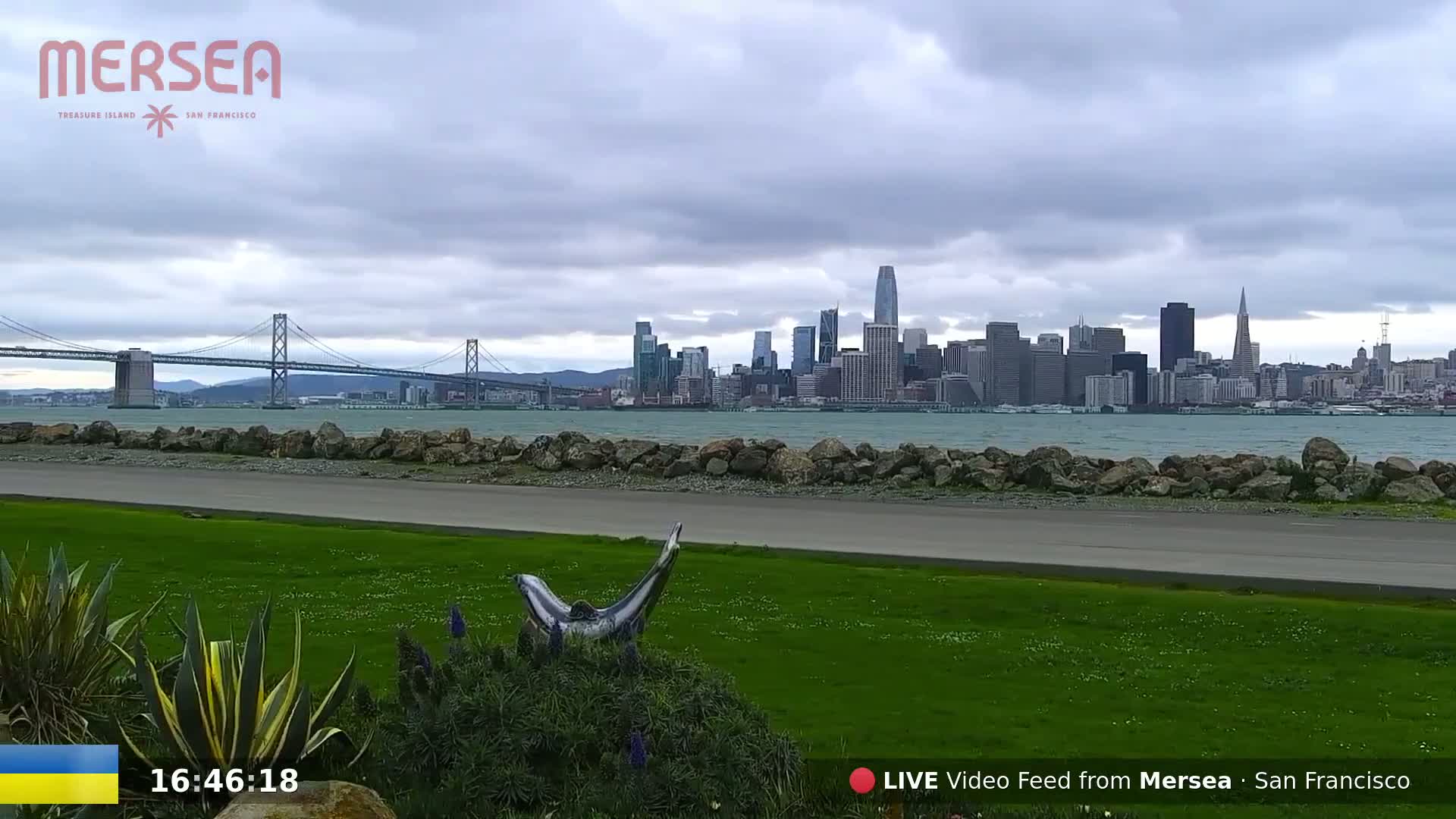 A grassy area with a dolphin sculpture overlooks a body of water, a cityscape, and a bridge under a cloudy sky.