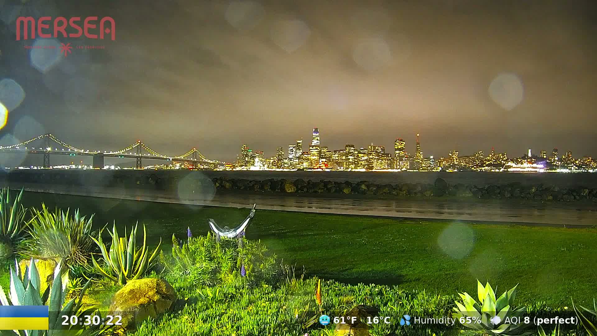 A nighttime view of a city skyline across a body of water, under a dark, rainy sky, with lush green landscaping in the foreground.